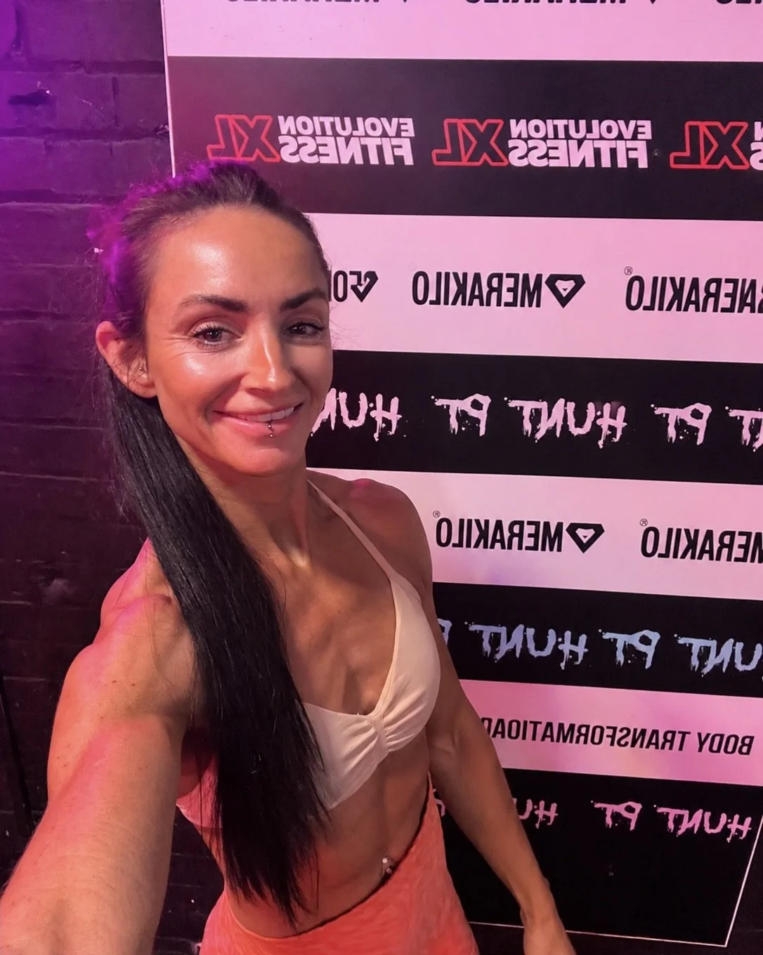 A woman with long dark hair, a piercing on her lip, and a toned physique, taking a selfie at a fitness event with a banner in the background. The banner reads 'Evolution Fitness' and 'PT HUNT'.