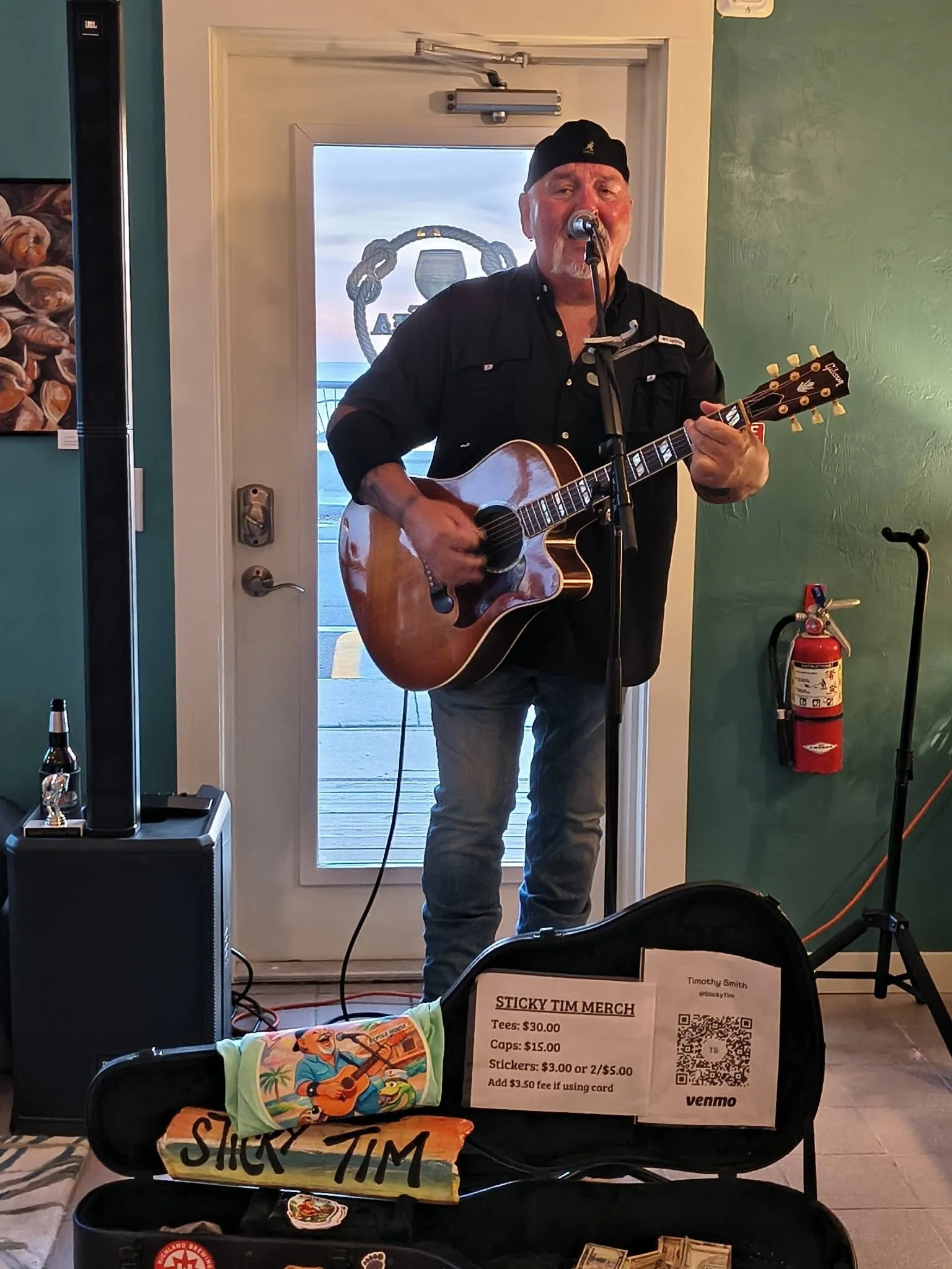A man singing and playing guitar in a room with green walls, in front of a door with a view of the gulf. In the foreground, a black case with with sticker reading 'STICKY TIM' holds cash, tee-shirts, caps and stickers for sale.
