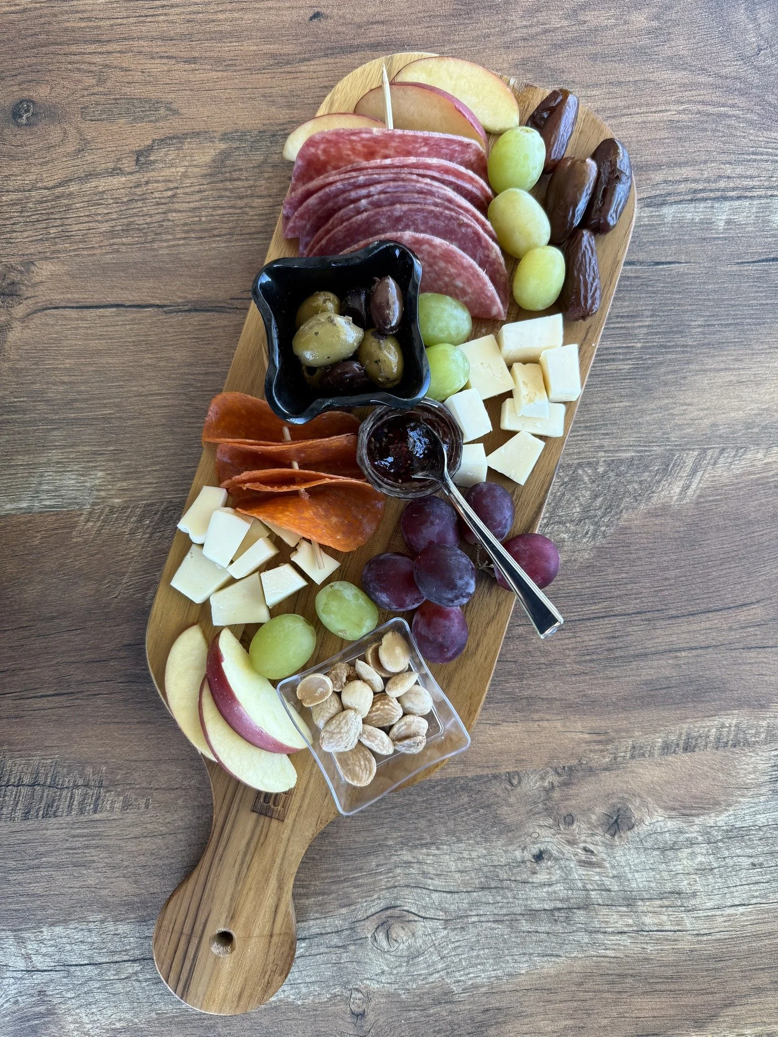 A cheese and charcuterie board, consisting of an assortment of sliced meats, cheeses, fresh fruit, nuts, olives and preserves on a long wooden cutting board.