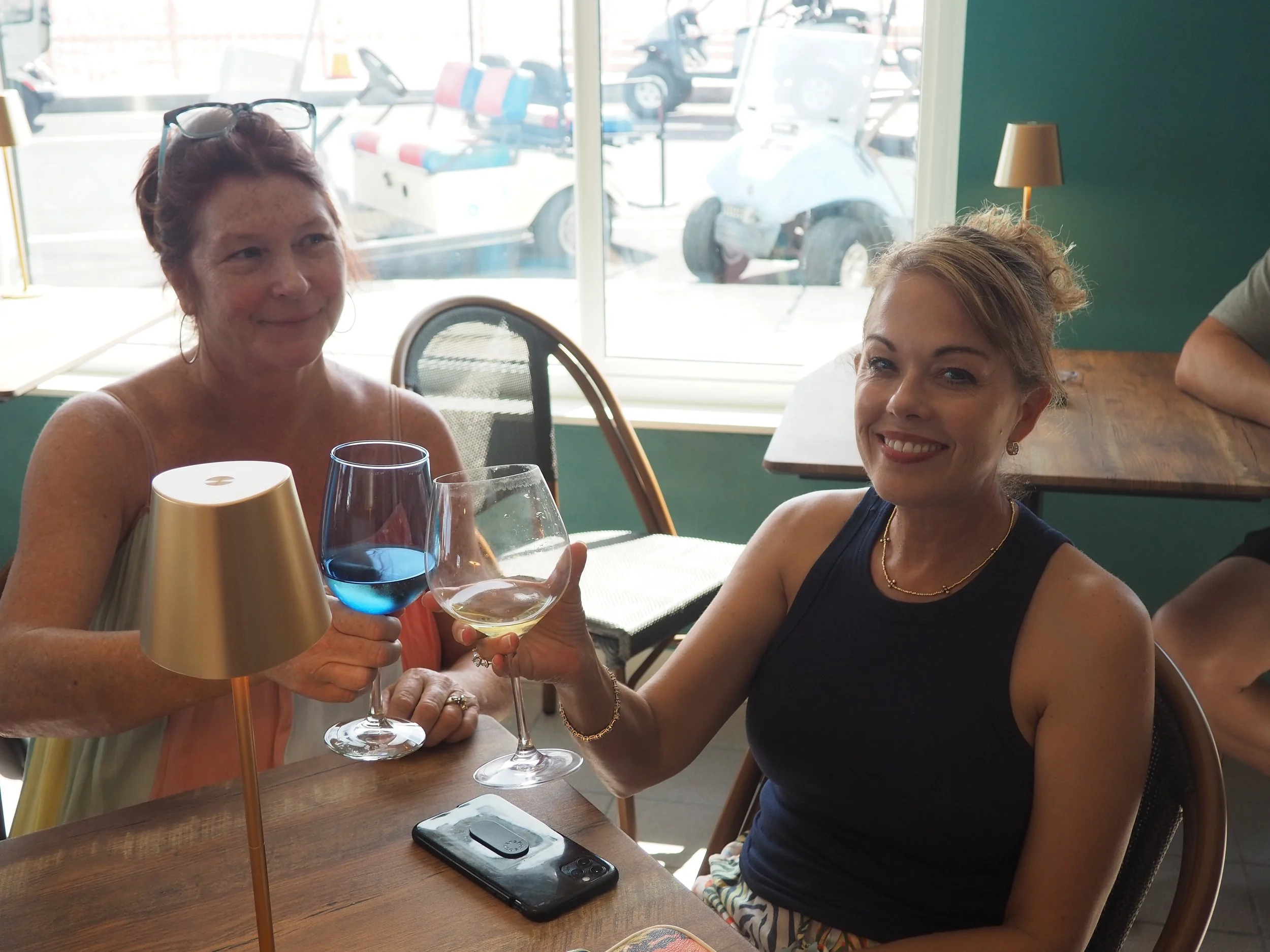 Two women sitting at a wooden table inside a cafe, toasting with wine glasses. One woman is wearing a sleeveless shirt and the other is holding her glasses. There is a mobile phone on the table and a gold lamp. Outside the window, parked cars and a g