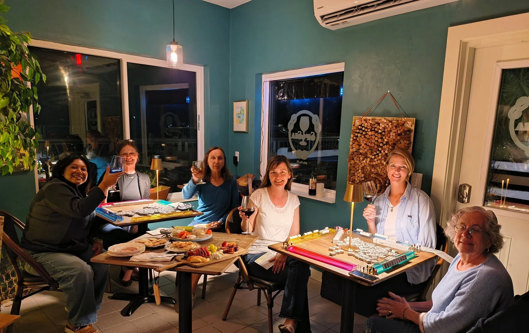 A group of people gathered around a dining table in a cozy, brightly lit room, celebrating with drinks and snacks, with some smiling and raising glasses. The room has green walls, large windows, and decorations including a firewood display and artwor