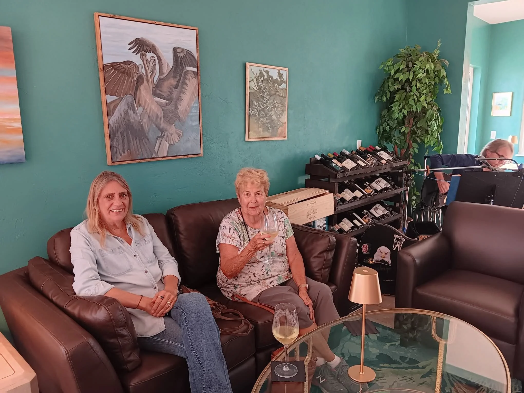 Two women sitting on a brown leather sofa with drinks in a cozy room with teal walls, colorful artwork, a wine rack, and a man behind a music stand near an open door.
