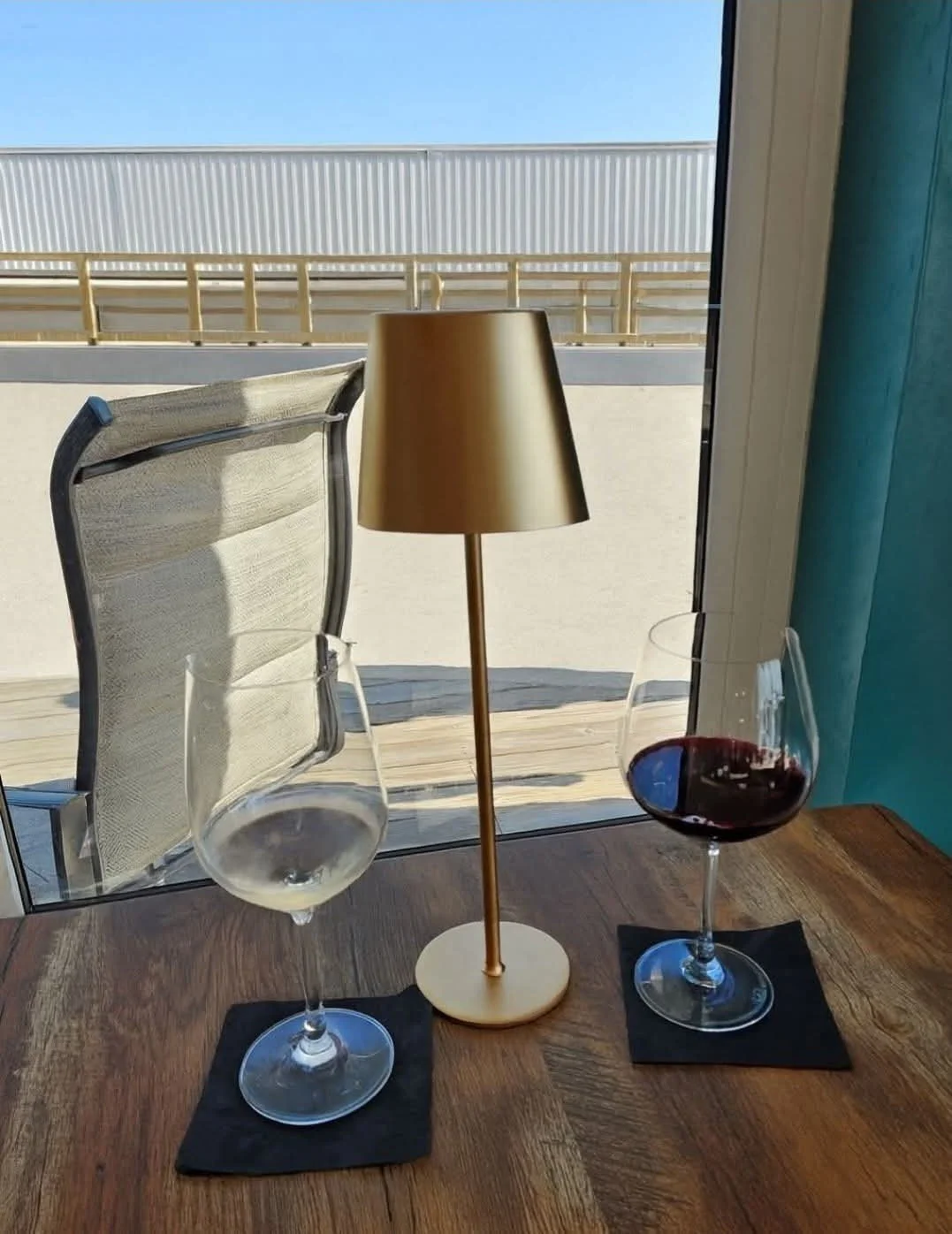 A wooden table with two wine glasses, one filled with white wine and the other with red wine, placed on black napkins. A small gold-colored lamp sits between the glasses. The scene is set by a window with a view of a dock and clear blue sky outside.