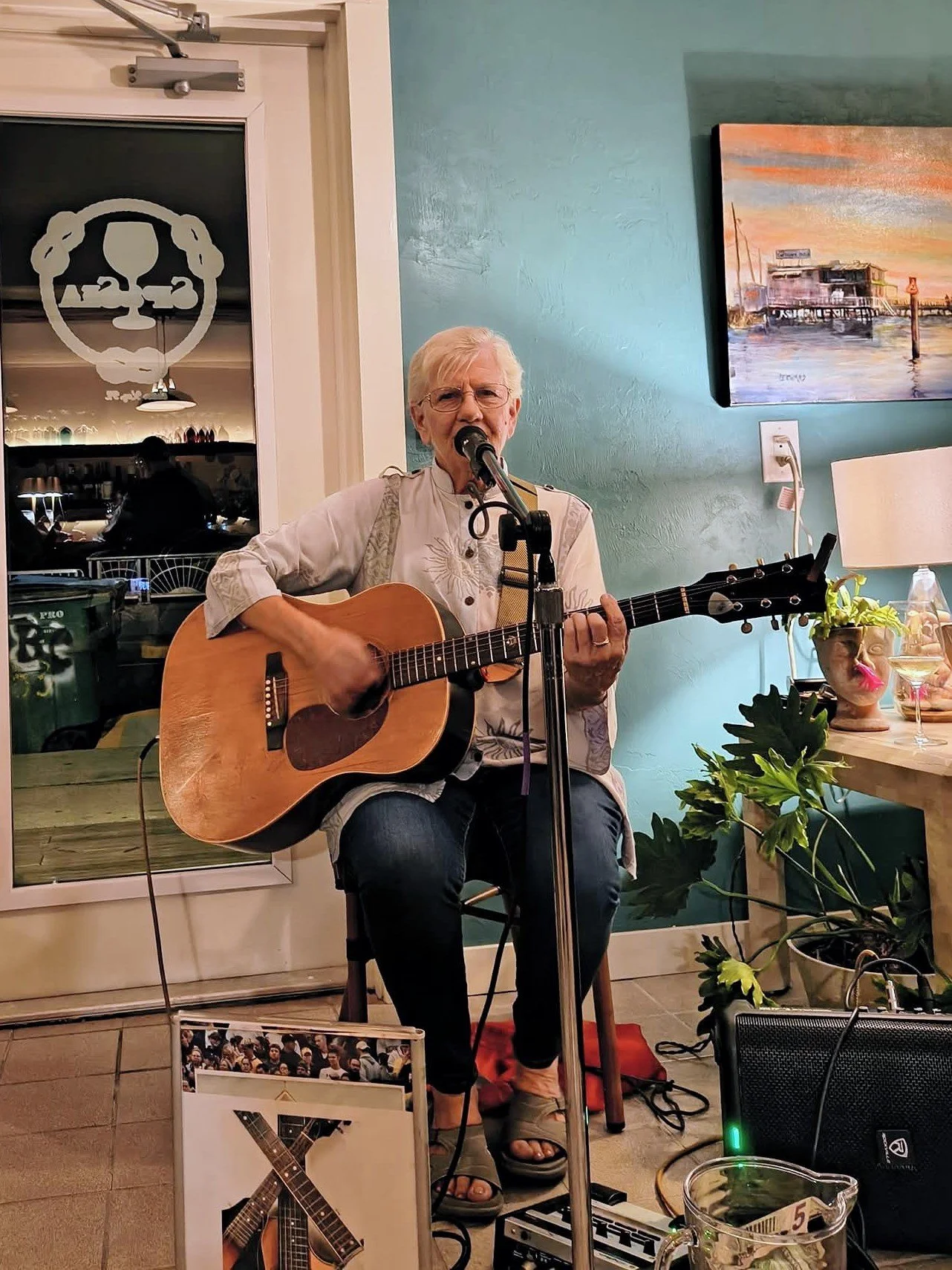An elderly woman with short gray hair and glasses is singing into a microphone while playing an acoustic guitar. She is sitting on a wooden chair in a cozy indoor setting, decorated with plants and wall art. A photo of a guitar is displayed on an eas