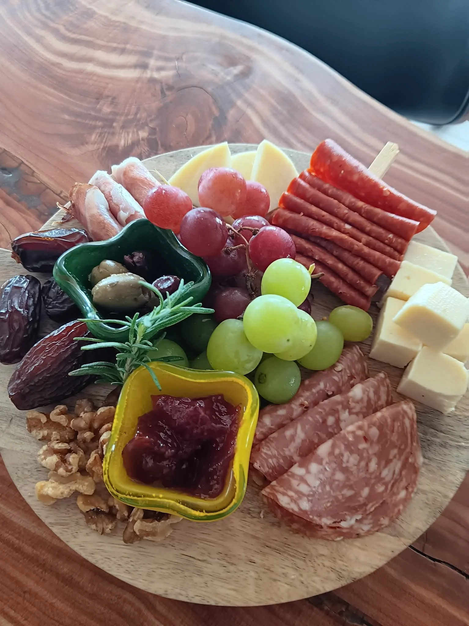 A round wooden serving board with assorted cheese, grapes, sliced meats, olives, jam, walnuts, and garnished with rosemary.