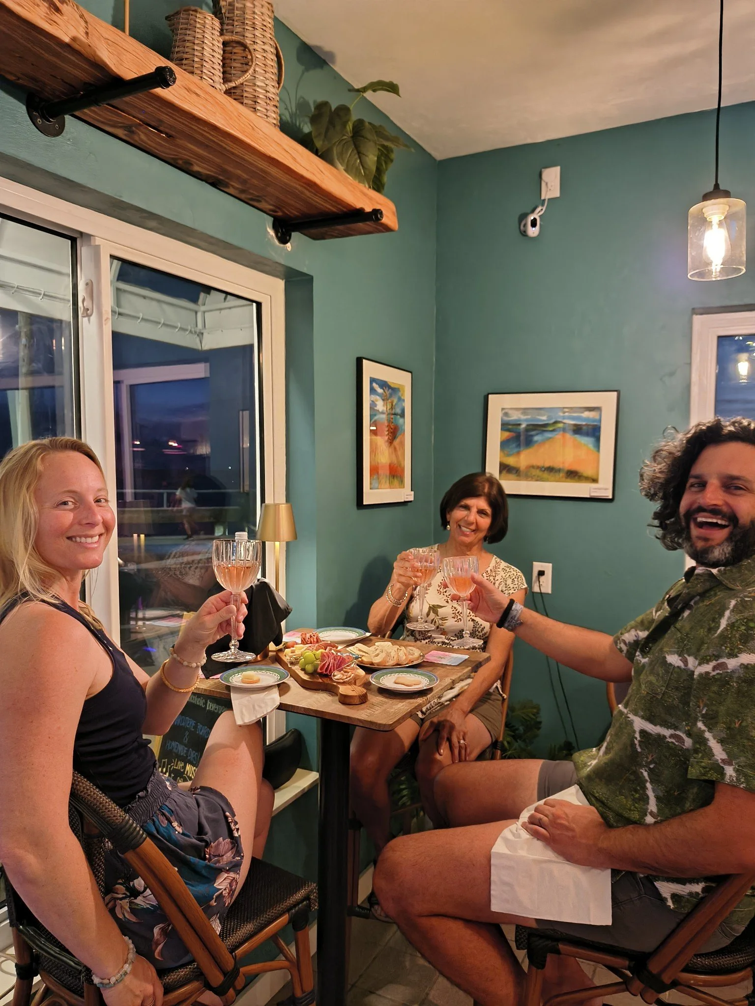 Three people celebrating with glasses of rosé wine in a cozy restaurant. A woman on the left wears a floral dress, another woman in the background has a short haircut, and a man on the right, with curly hair and a patterned shirt. A plate of charcute