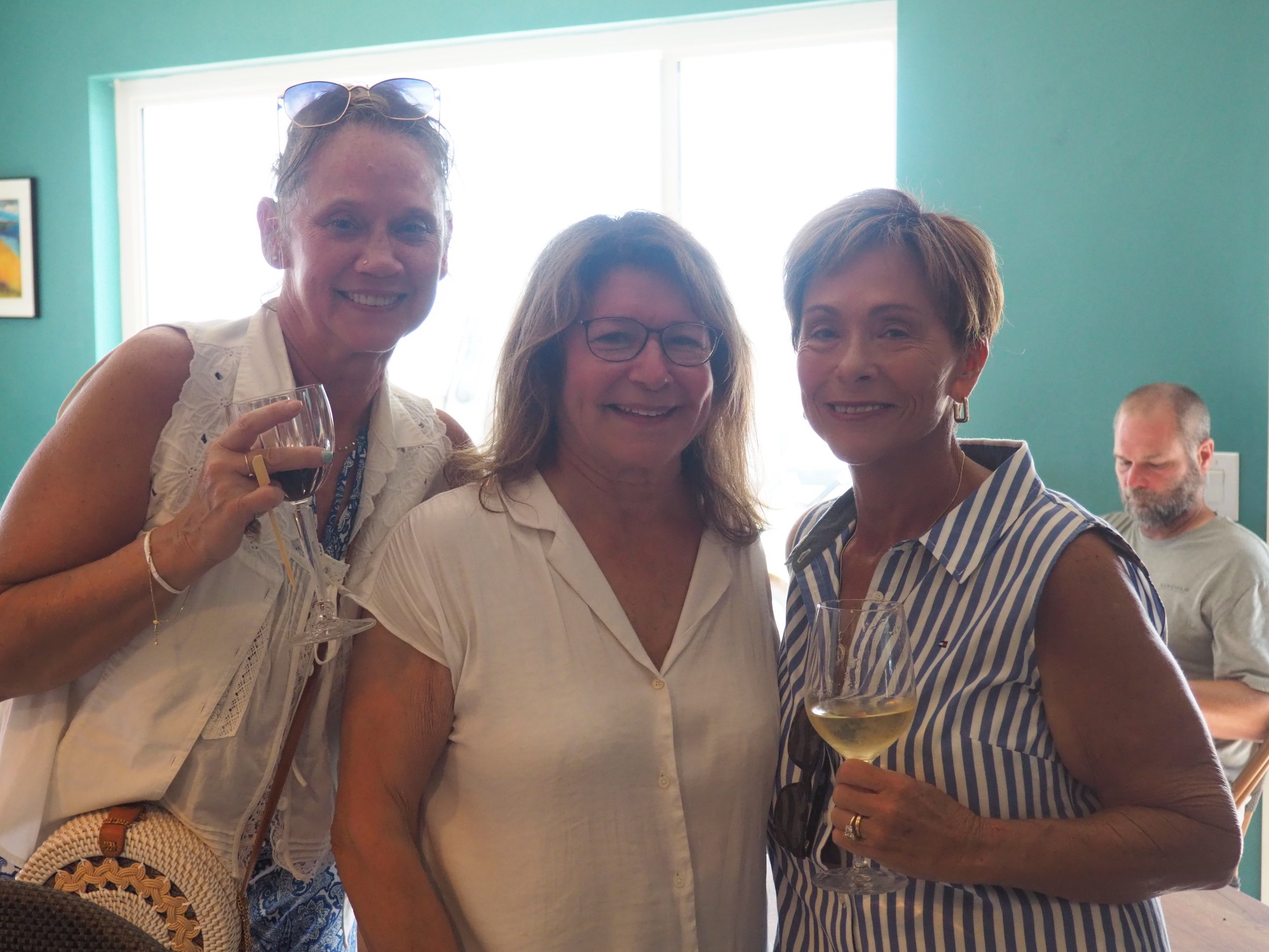 Three women smiling and holding drinks, two with wine glasses, in a brightly lit room with teal walls and artwork, and a man in the background sitting and looking down.