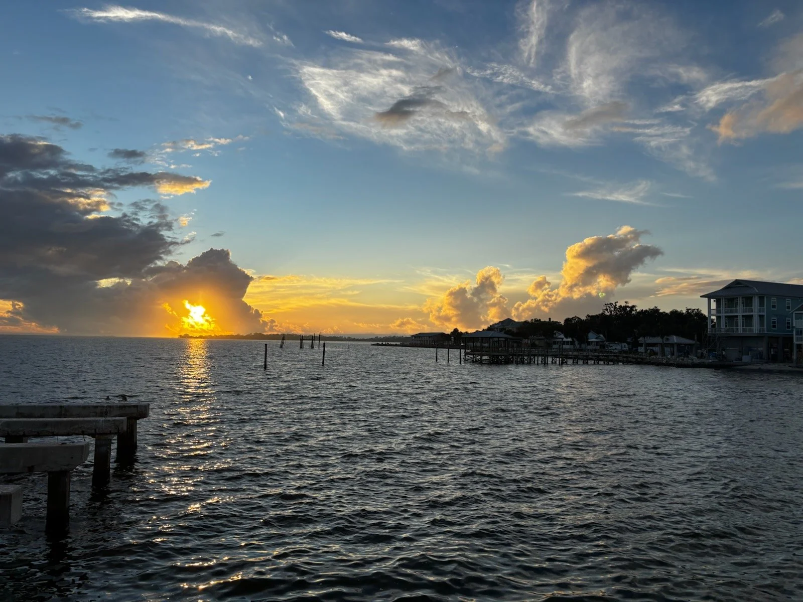 Sunset over a body of water with clouds and dock structures, residential buildings on the right, and a partially cloudy sky.
