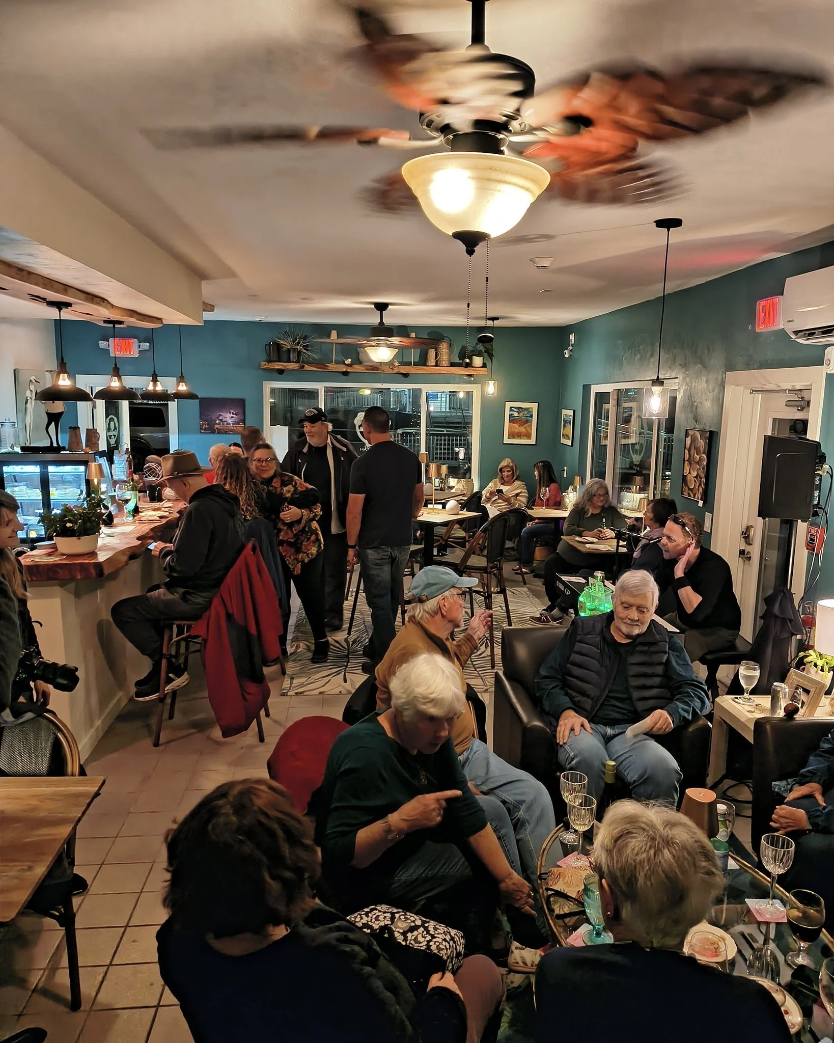 A crowded restaurant with people sitting at tables, some using phones, having drinks and conversations. A ceiling fan is spinning, creating a blurred motion effect. The walls are decorated with framed pictures, and there is a counter area with cream-