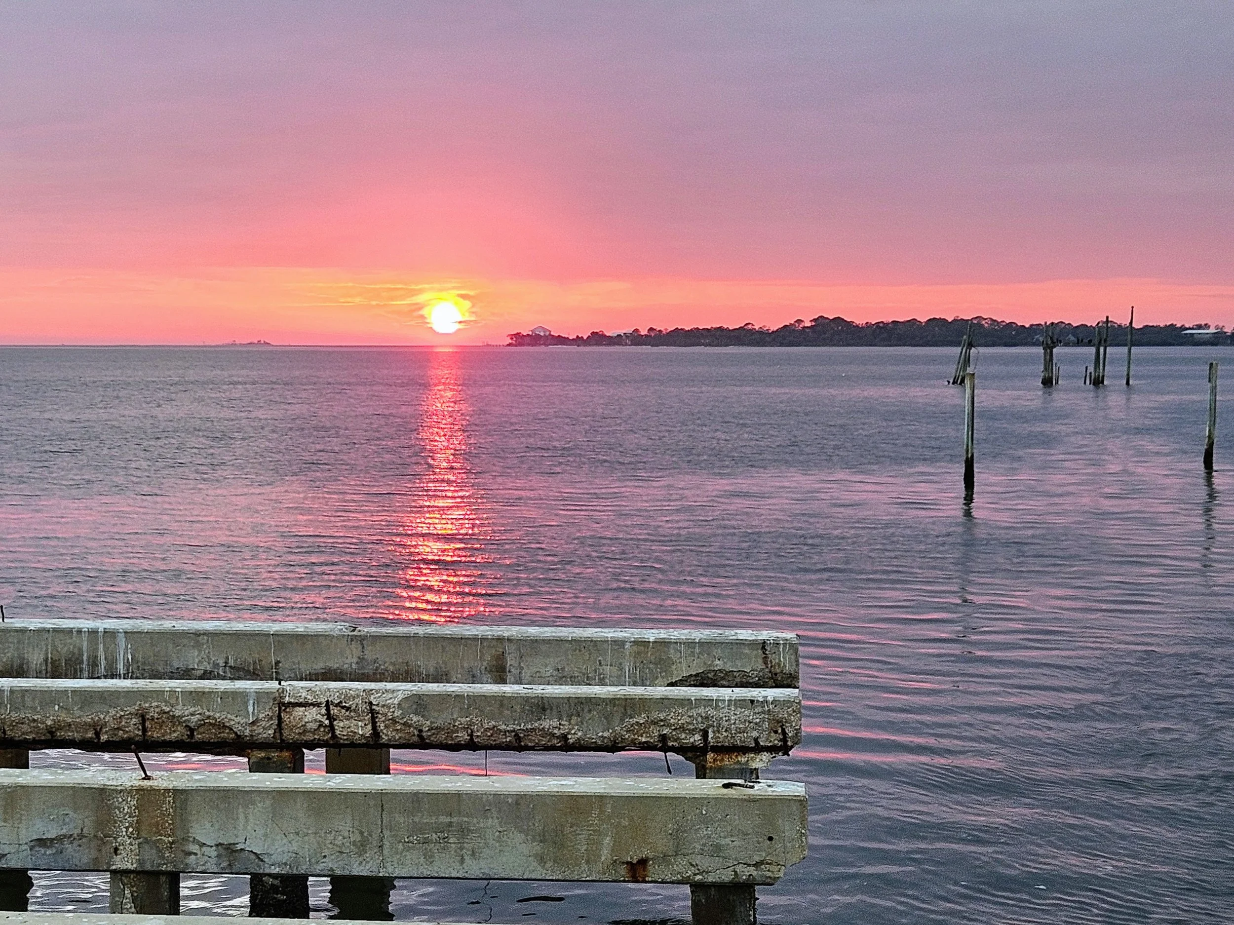 Sunset over calm water with pink and purple sky, reflections on the water, weathered wooden dock and posts in the foreground.