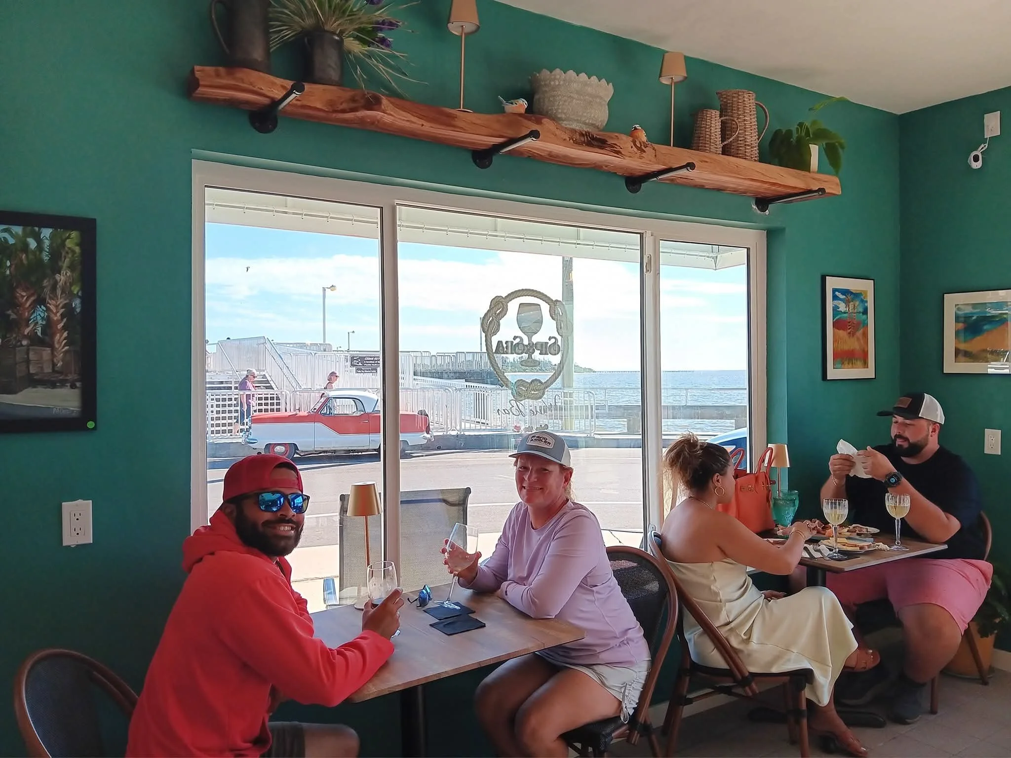 Four people sitting at tables inside a restaurant with a view of the outside water and dock, enjoying drinks and food