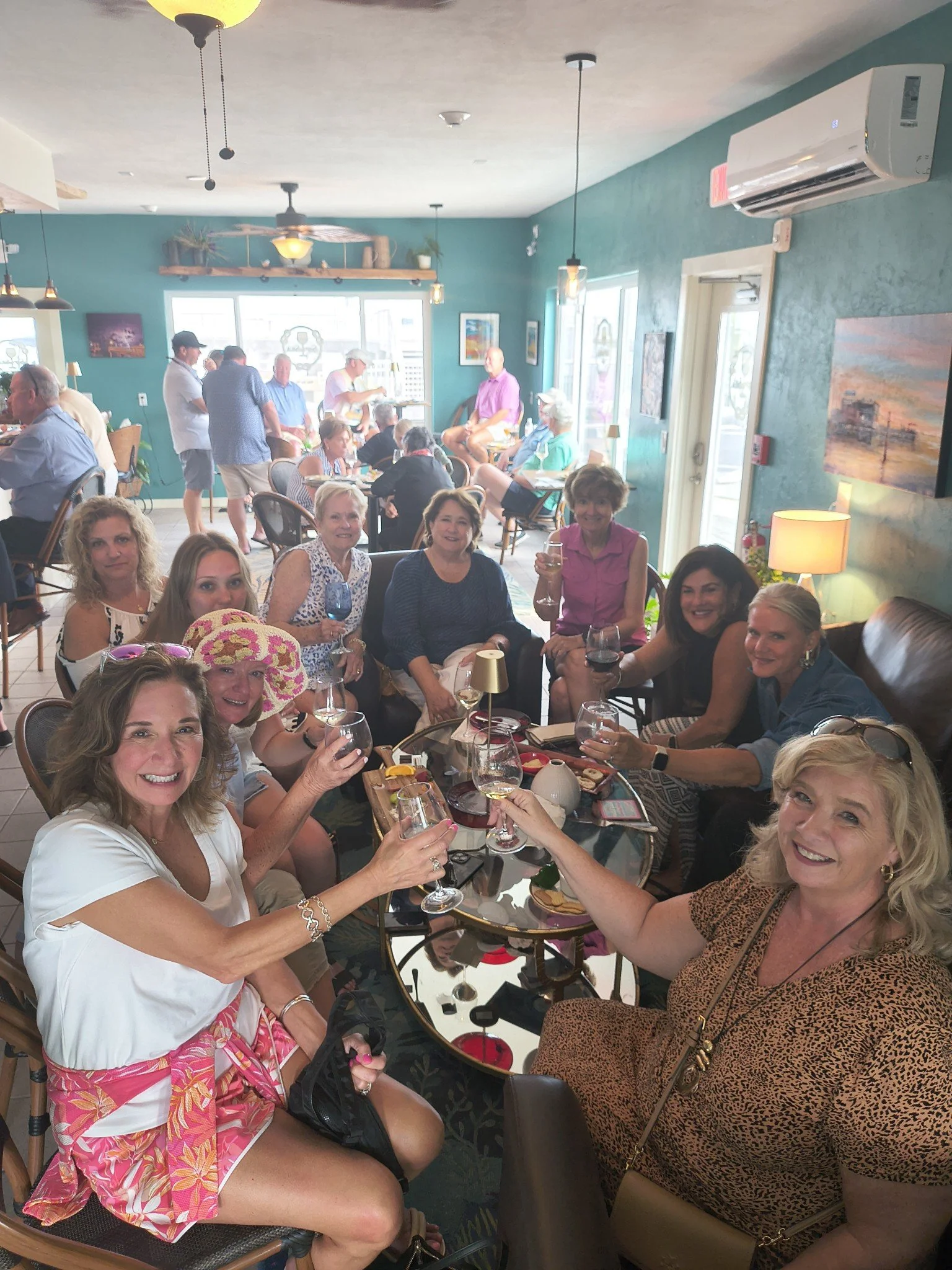 A group of women sitting at a table in a restaurant, smiling and holding wine glasses, with other patrons and decorations visible in the background.
