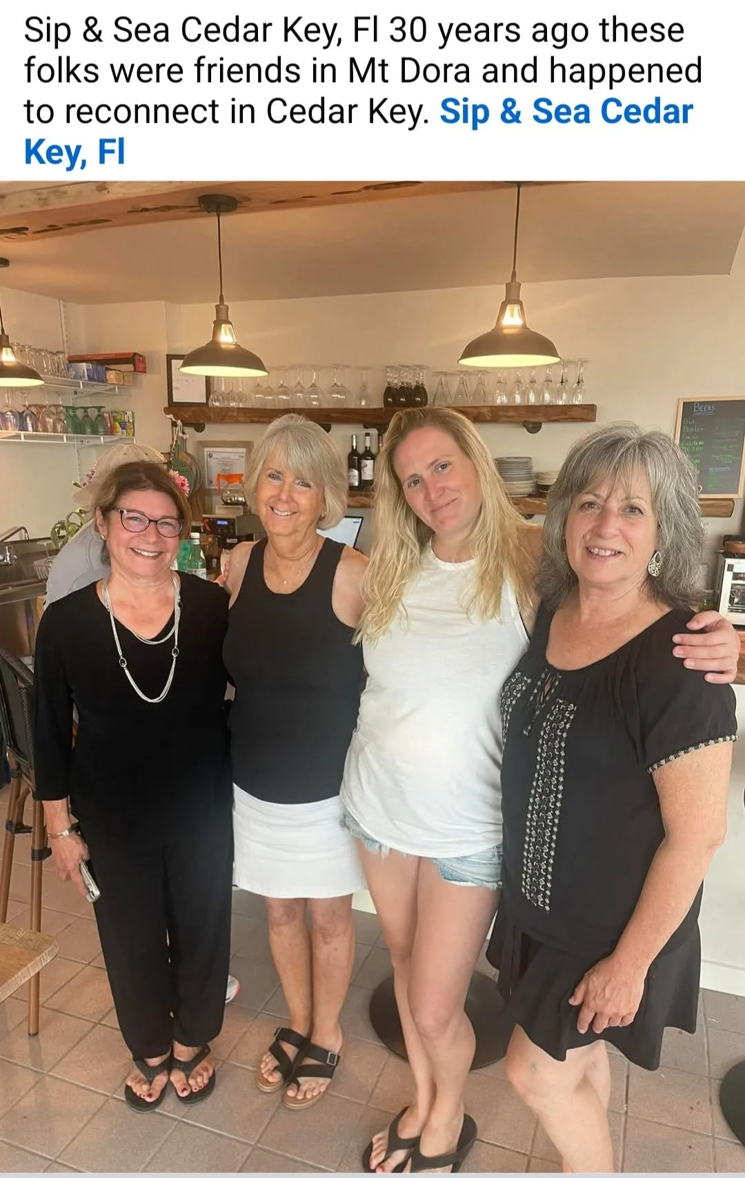 Four women standing together in a restaurant kitchen smiling at the camera, with shelves of glasses and bottles behind them.