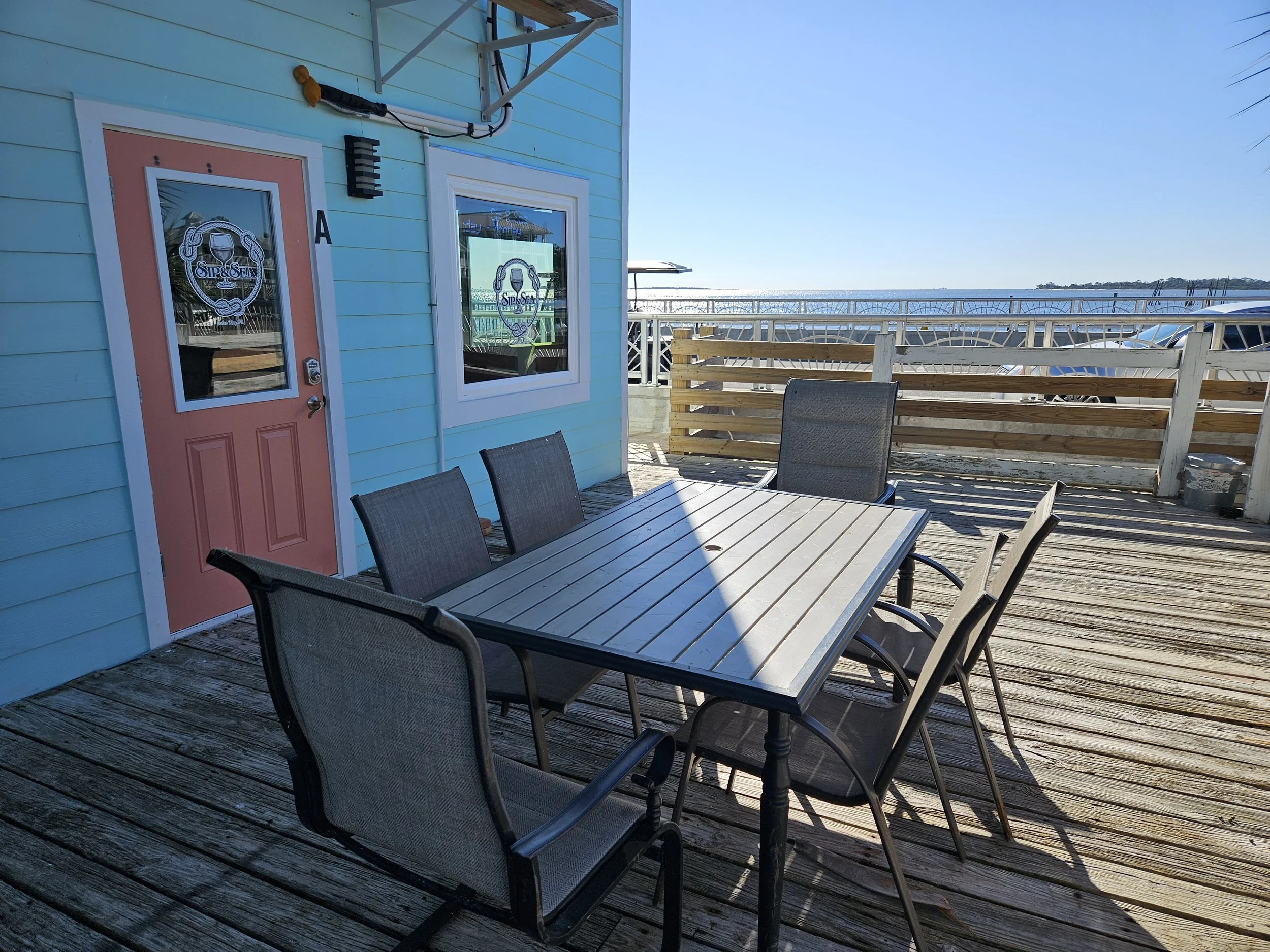 An outdoor patio with a wooden table and six chairs next to a light blue building with a pink door, overlooking the water and sky in the background.
