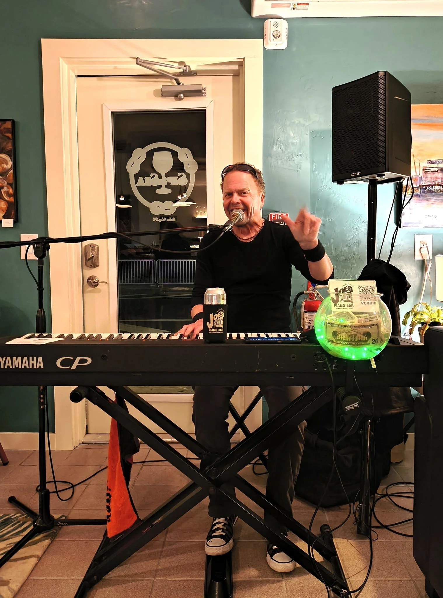 A man singing and playing keyboard at a music venue, with a microphone, speaker, and fishbowl with a paper and QR code on it.