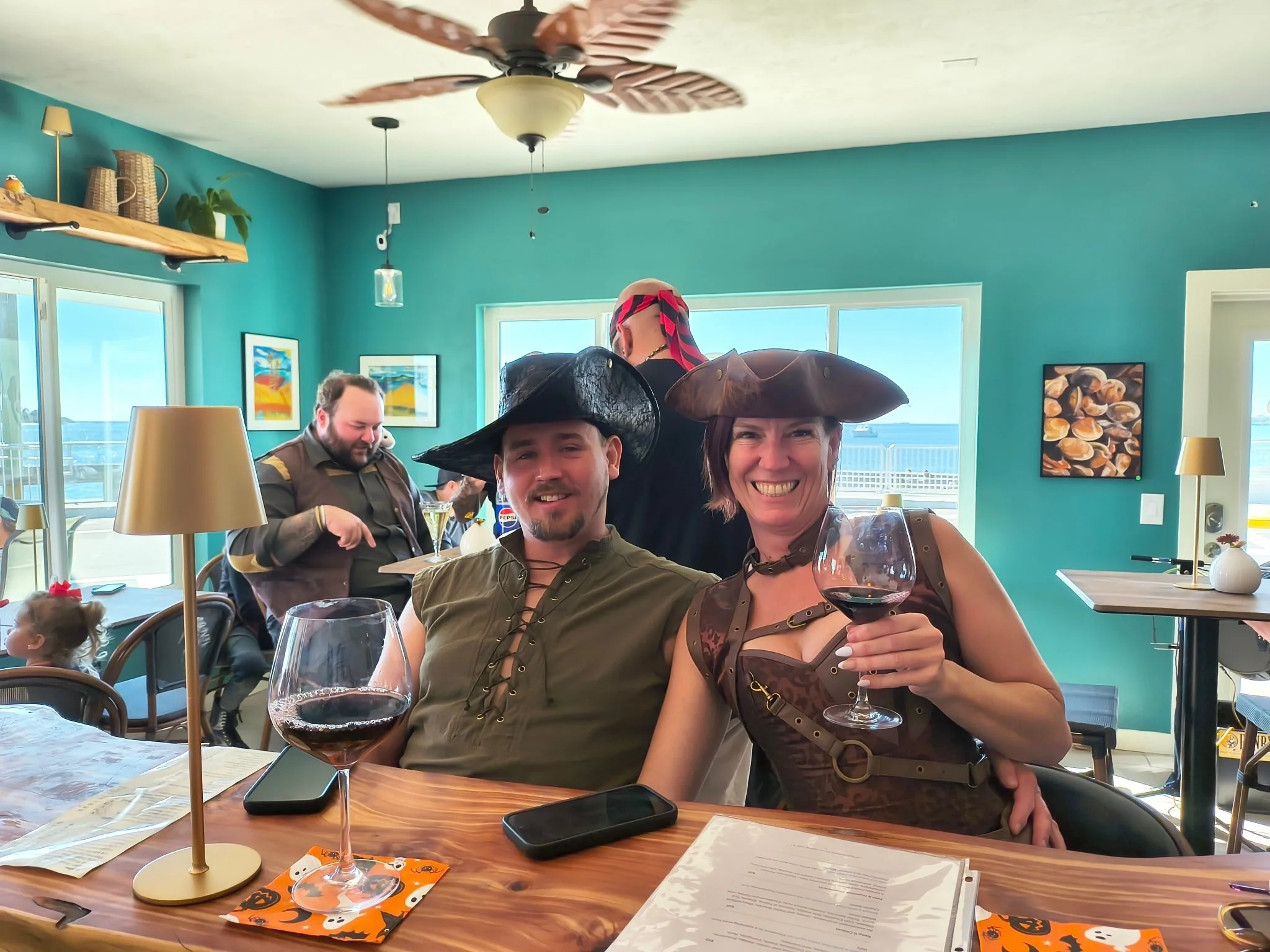 Group of people in a colorful restaurant wearing pirate costumes with hats, holding wine glasses, celebrating. Two women in the foreground are smiling and holding glasses of red wine, and there are other guests in the background. The room has windows