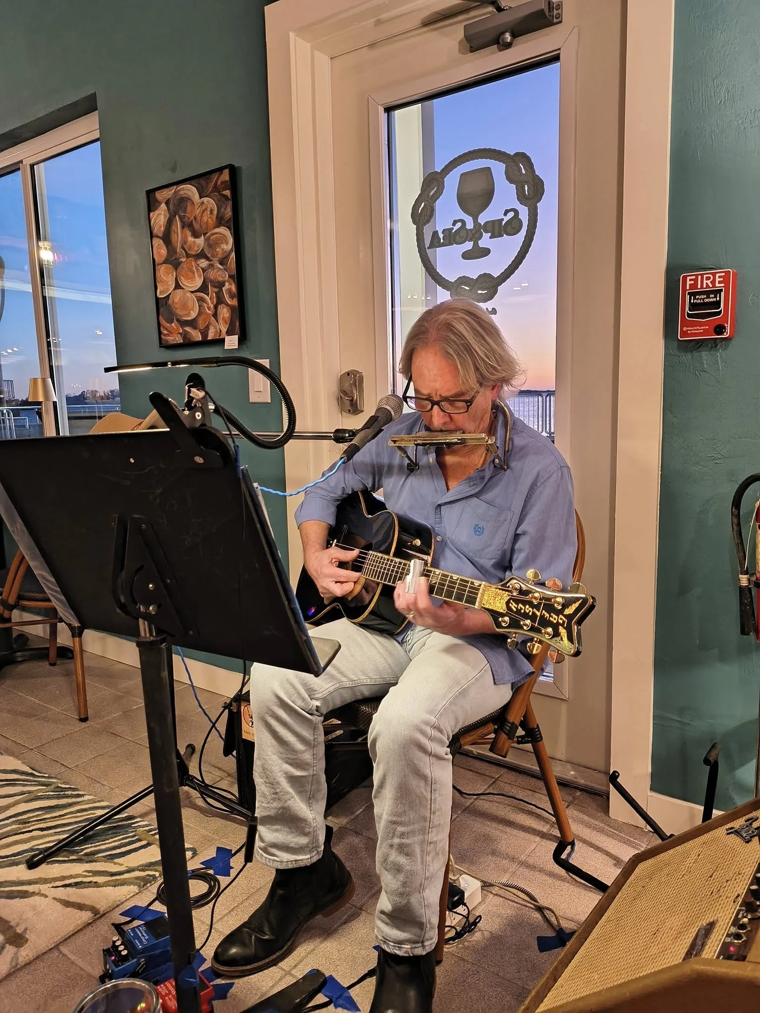 A man with gray hair and glasses playing an acoustic guitar while sitting on a chair, with a harmonica holder around his neck, in a room with teal walls and a window showing a sunset outside. There is a microphone in front of him and a music stand ne