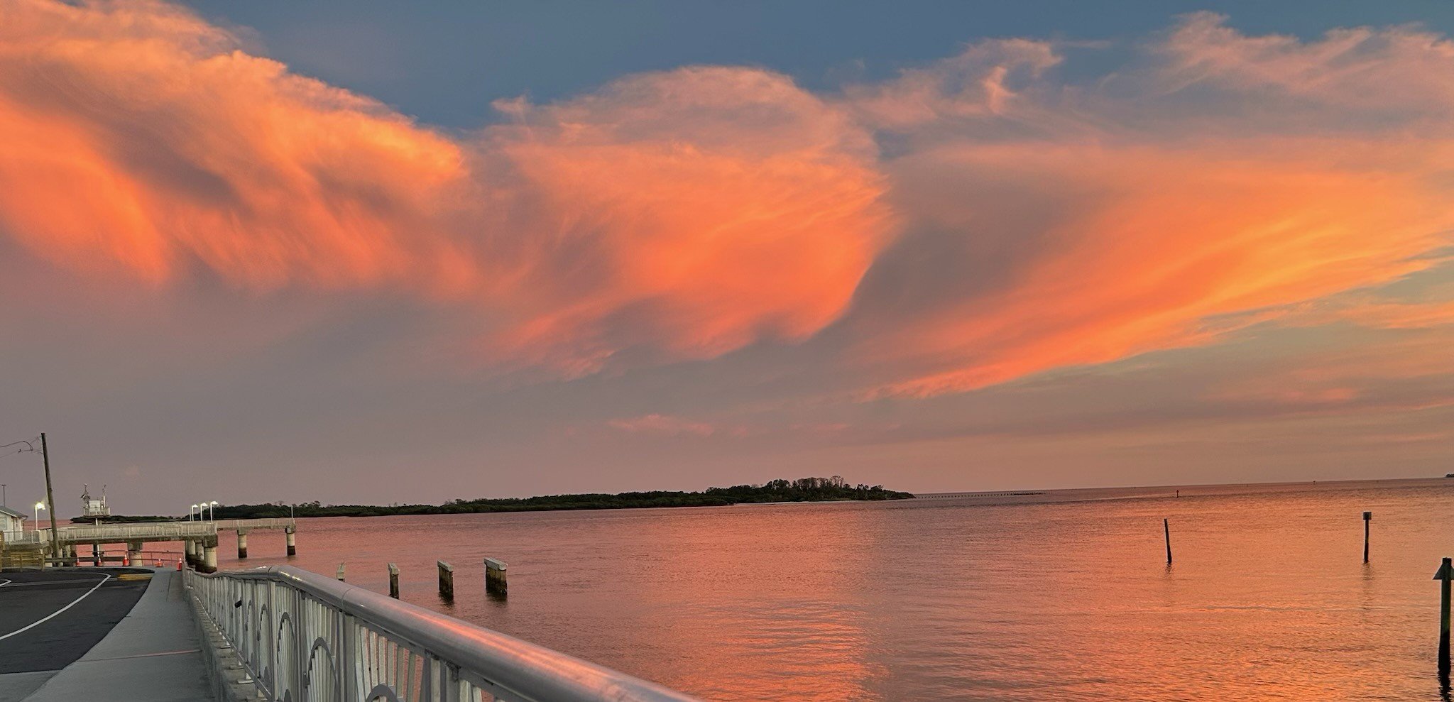 A sunset over the Gulf of Mexico with pink and orange clouds in the sky. There is a pier or dock with railings on the left side and a small island or landmass in the distance.