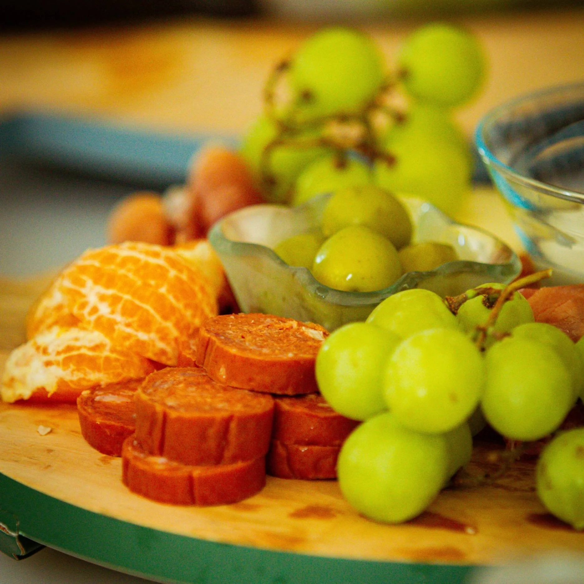 Close-up of a charcuterie board with slices of salami, green olives, clusters of green grapes, and peeled mandarin wedges.