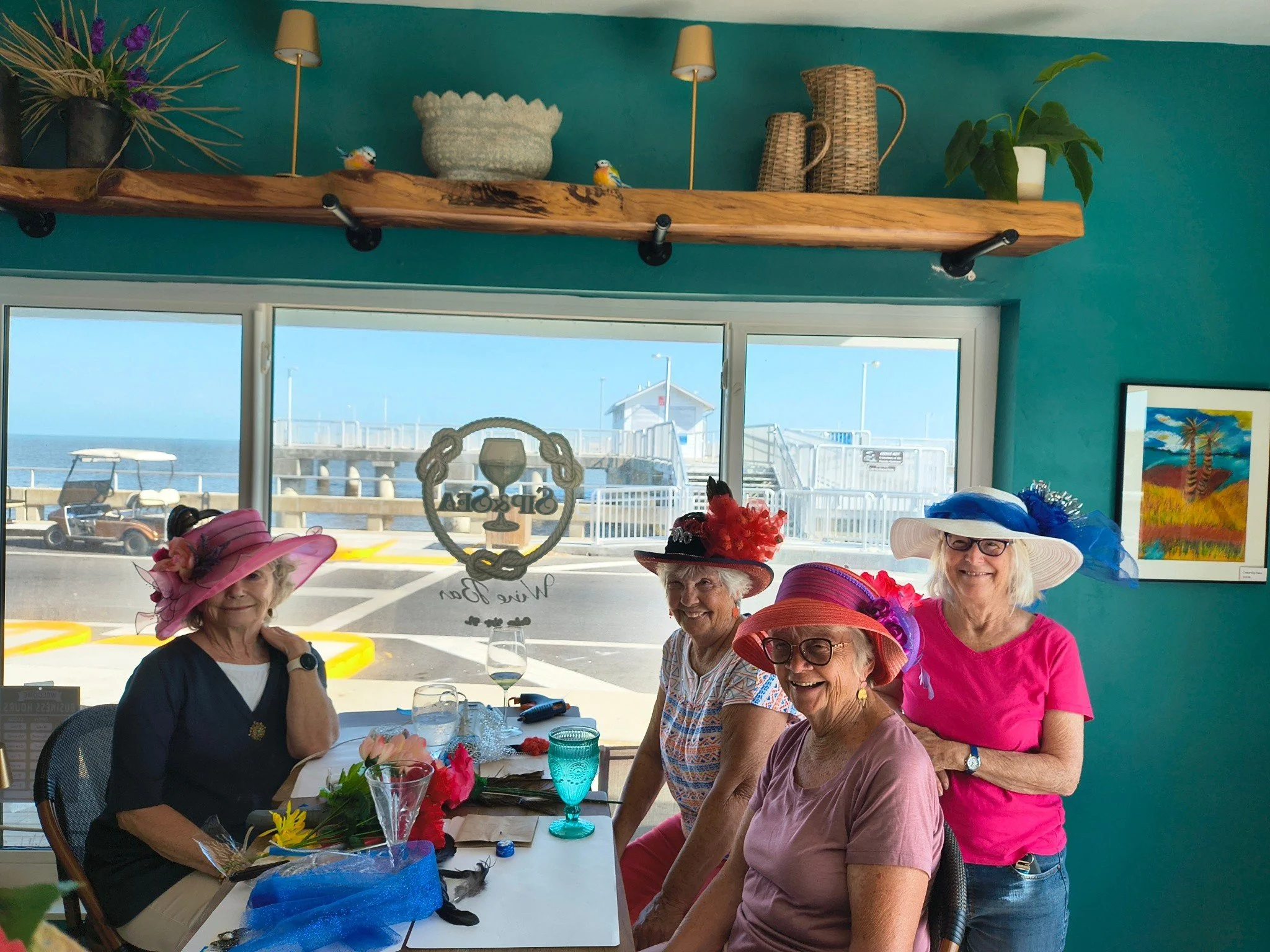 Group of older women enjoying a Derby Hat party in a wine bar with large hats, bright clothing, and colorful decorations, by a window showing a pier outside.