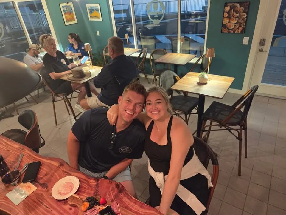 A couple smiling and posing for a photo in a restaurant. The man has short hair and is wearing a dark shirt. The woman has blonde hair and is wearing a black top. On the table, there are drinks, sunglasses, a phone, and a dessert plate. In the backgr