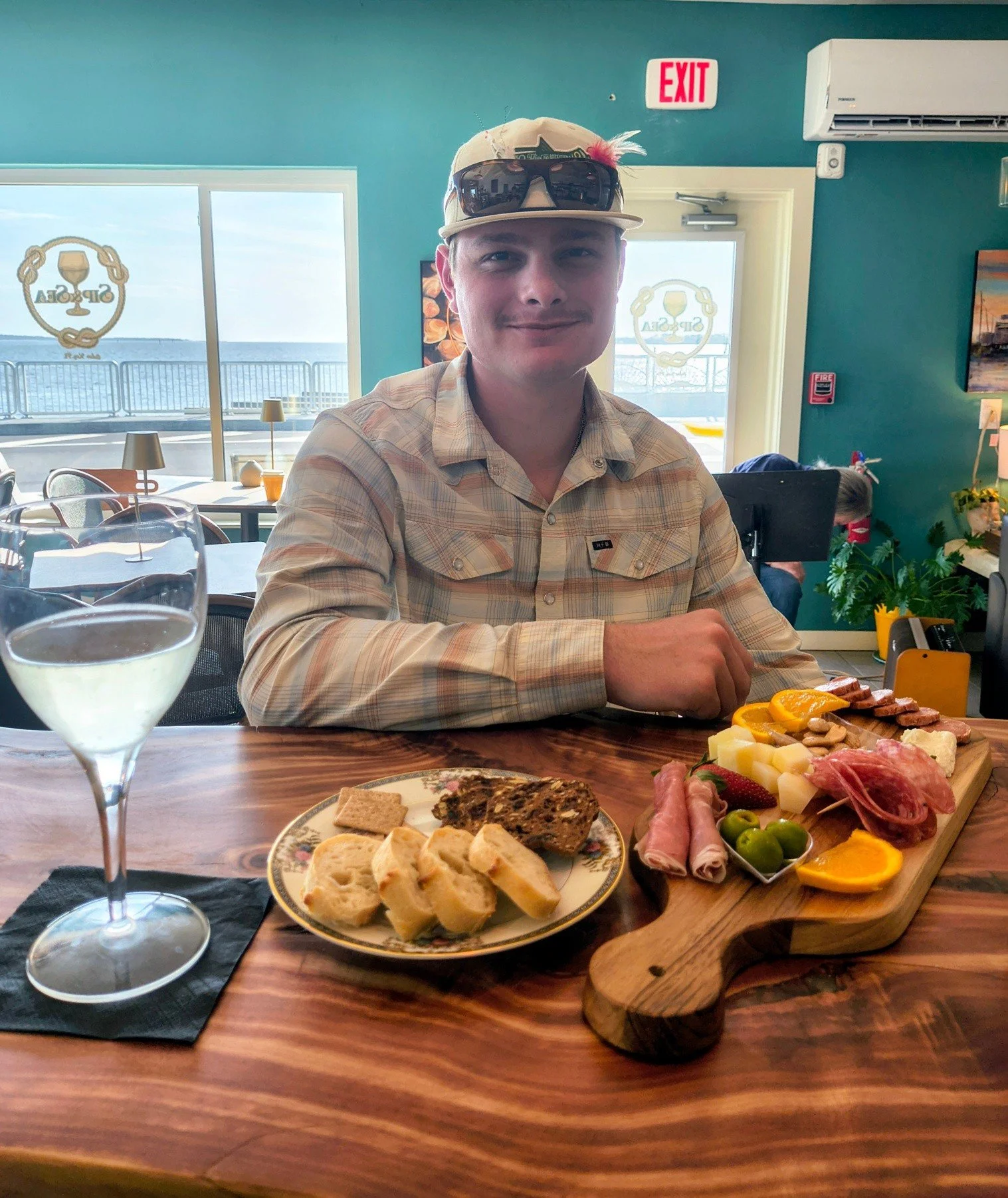 A man with a beige cap and sunglasses on top, sitting at a wooden bar top with a charcuterie board, a plate of crackers and sliced baguette, and a glass of white wine, with a gulf view through large windows in the background.