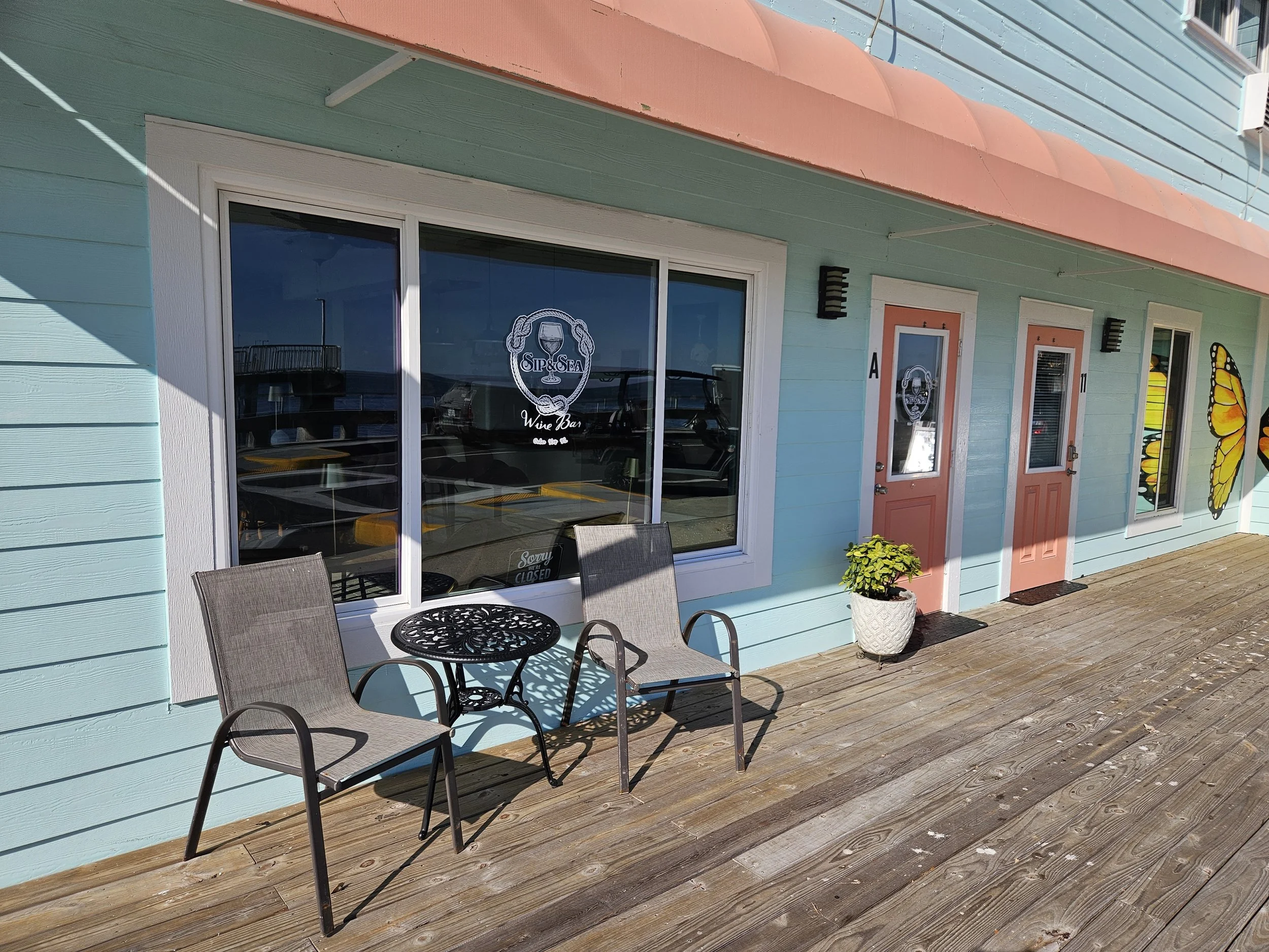 Exterior of a pastel blue building with pink doors and mural artwork of butterfly wings. There are two chairs and a small table on the wooden deck in front of a large window with a sign that says 'Sip & Sea Wine Bar.'