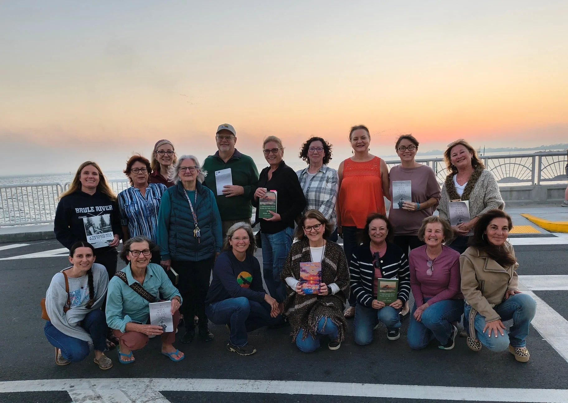 Group of women and one man posing on a street near the water at sunset, holding books and smiling.