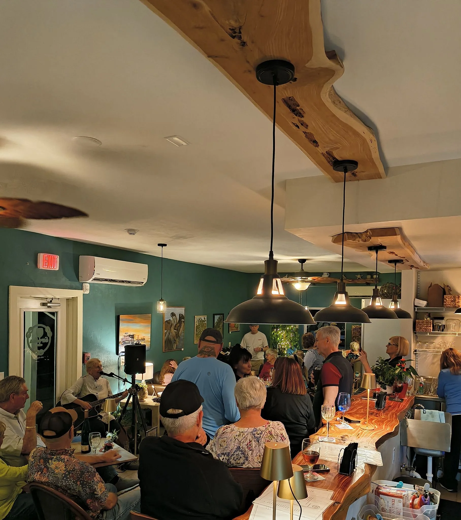 A lively indoor gathering with people socializing, some sitting at tables and others standing, many with drinks. A woman is playing a guitar and singing, while others are enjoying the atmosphere. The room has warm lighting, wooden accents, and severa