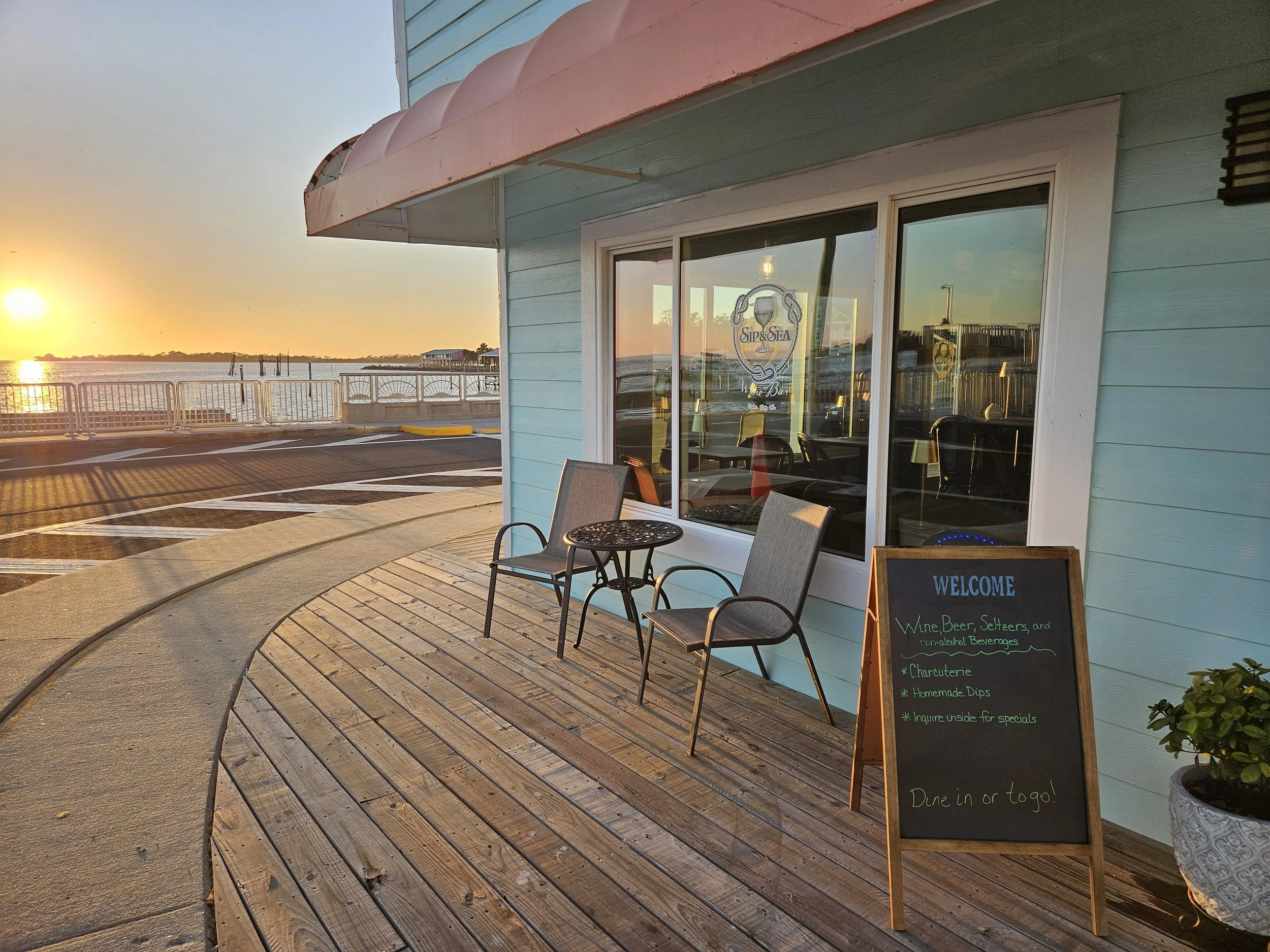Exterior of a waterfront restaurant or cafe with two chairs and a small table on a wooden deck, a blackboard sign advertising wine, beer, seltzers, charcuterie, and homemade dips, with a sunset view over the water and a parking lot in the background.