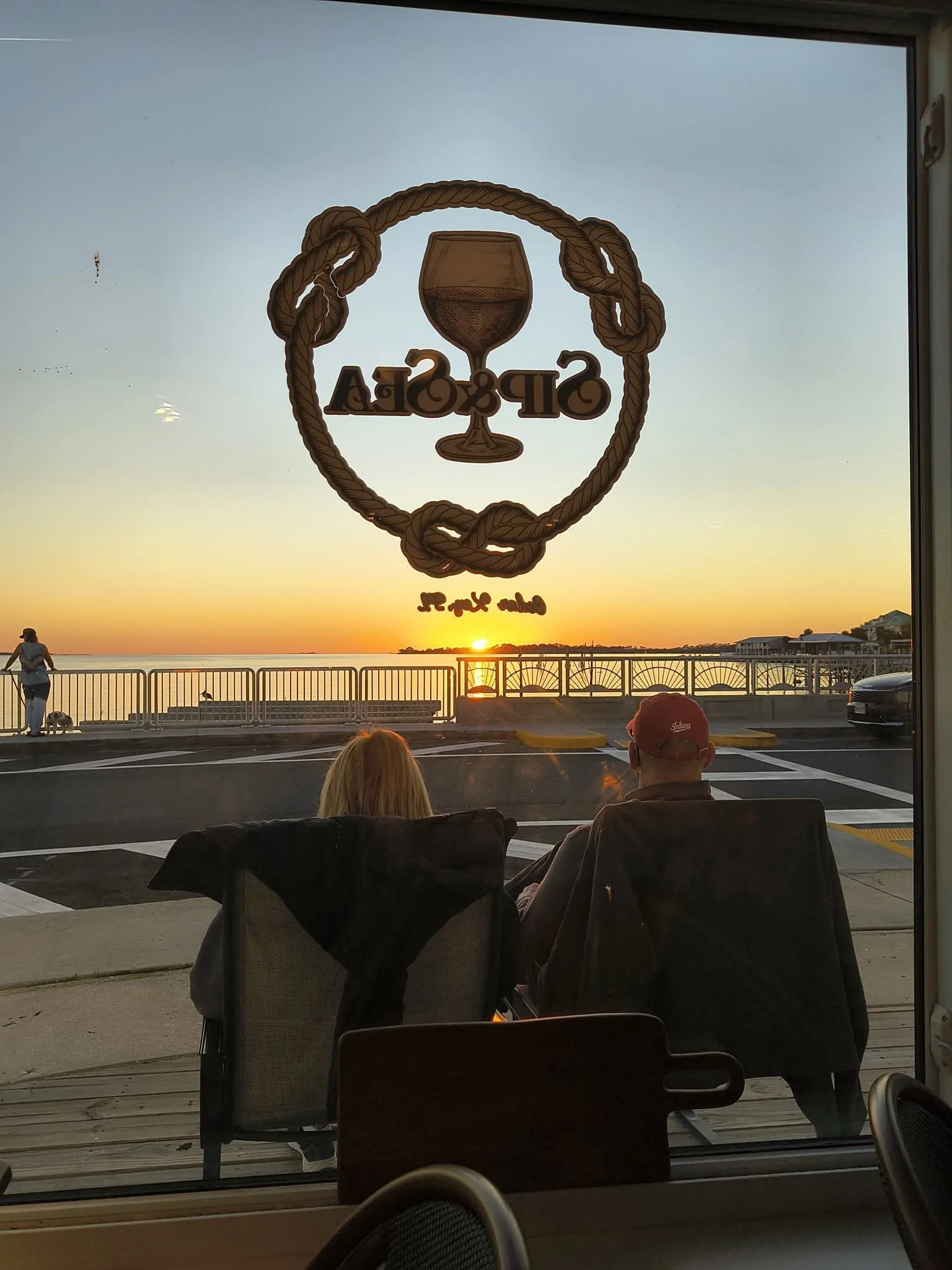 Visible through a window, two people sitting in chairs outside Sip & Sea Wine Bar looking at a sunset over the Gulf of Mexico.