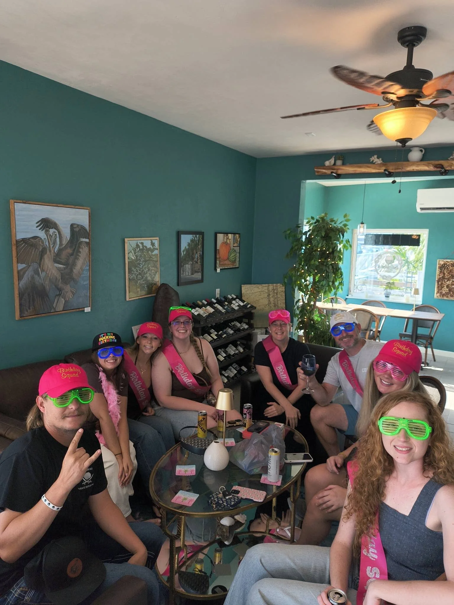 A group of people celebrating a birthday party in a wine bar, wearing pink birthday sashes and hats, with some wearing colorful sunglasses, sitting in couches around a glass table with drinks, candles, and party decorations, smiling at the camera.