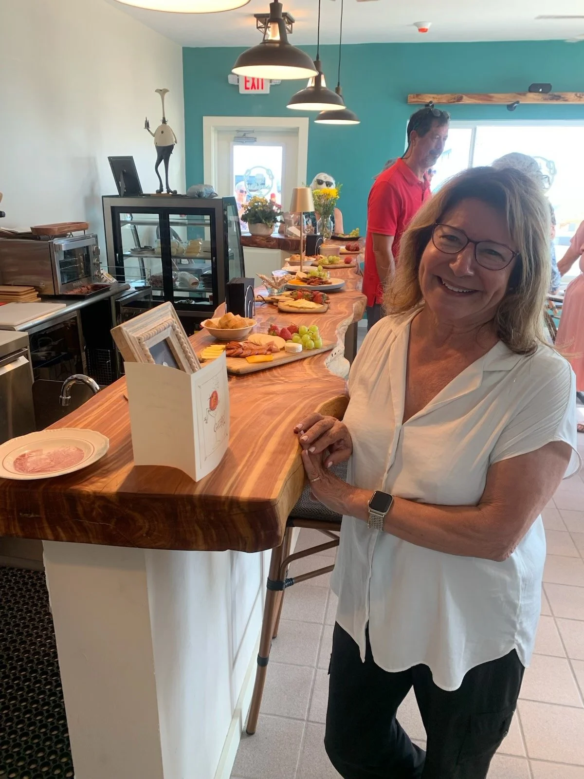 Smiling woman with glasses and a white blouse standing at a wooden counter with a spread of food and cheese, in a bright restaurant or cafe with teal walls and hanging lamps.