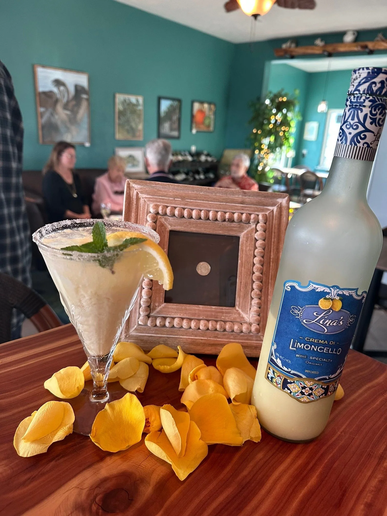A cocktail with a lemon and mint garnish in a glass, yellow flower petals scattered on a wooden table, a wooden picture frame, a bottle of limoncello, and a blurred background of people in a restaurant with teal walls and artwork.