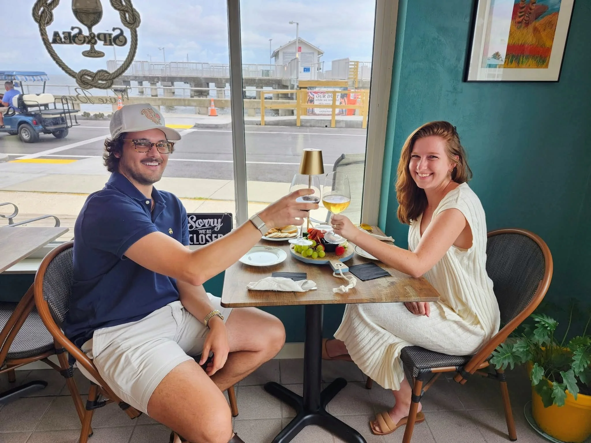 A man and woman smiling and toasting with drinks at a restaurant table near a window with a view of a beach and boardwalk outside; the table has a fruit platter, wine glasses, and plates.