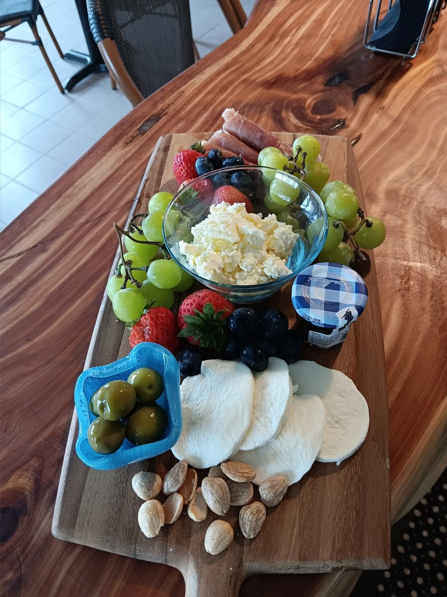 Assorted cheeses, green and red grapes, strawberries, blueberries, blackberries, green olives, chocolate hazelnut spread, ham, whipped cheese, and almonds on a wooden serving board.