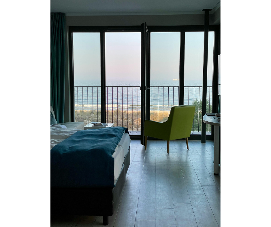 Room with a large glass door leading to a balcony overlooking the ocean, with a bed on the left, a green armchair near the door, and a small desk on the right.