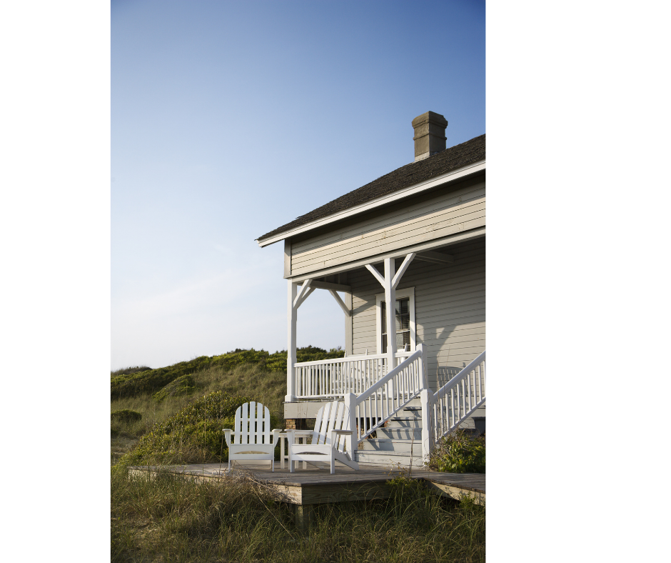 A small, two-story house with a white wooden exterior, a front porch with white railing, and Adirondack chairs on a grassy area in front. The house has a chimney, and the scene appears peaceful with a clear sky in the background.