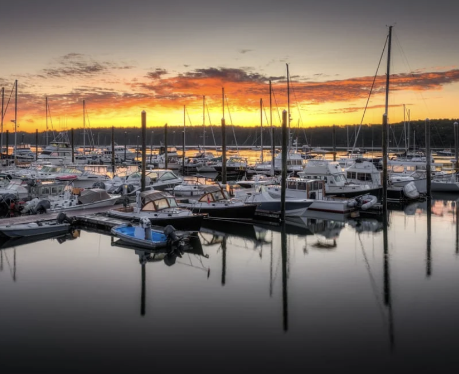 Marina with boats docked at sunset, colorful sky with orange and yellow hues, calm water reflecting boats and sunset