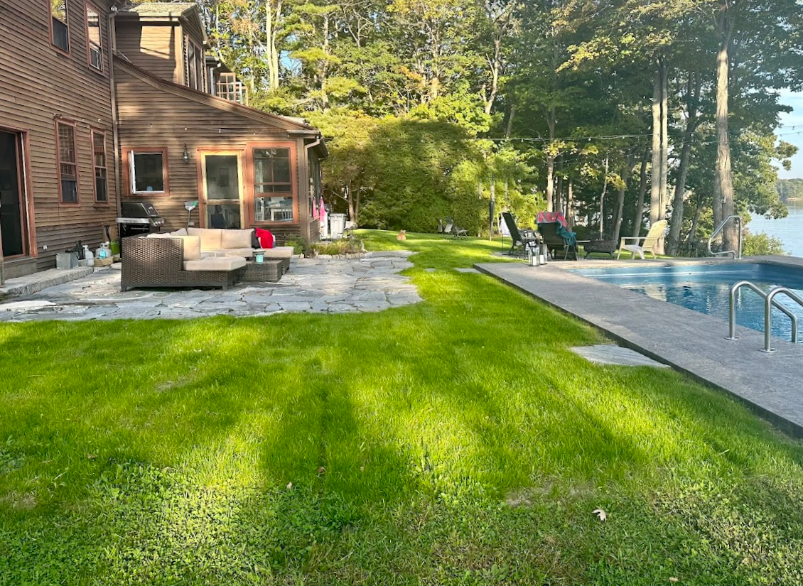 A backyard with a well-maintained green lawn, a stone patio with outdoor furniture, and a swimming pool with a view of trees and the ocean in the background.