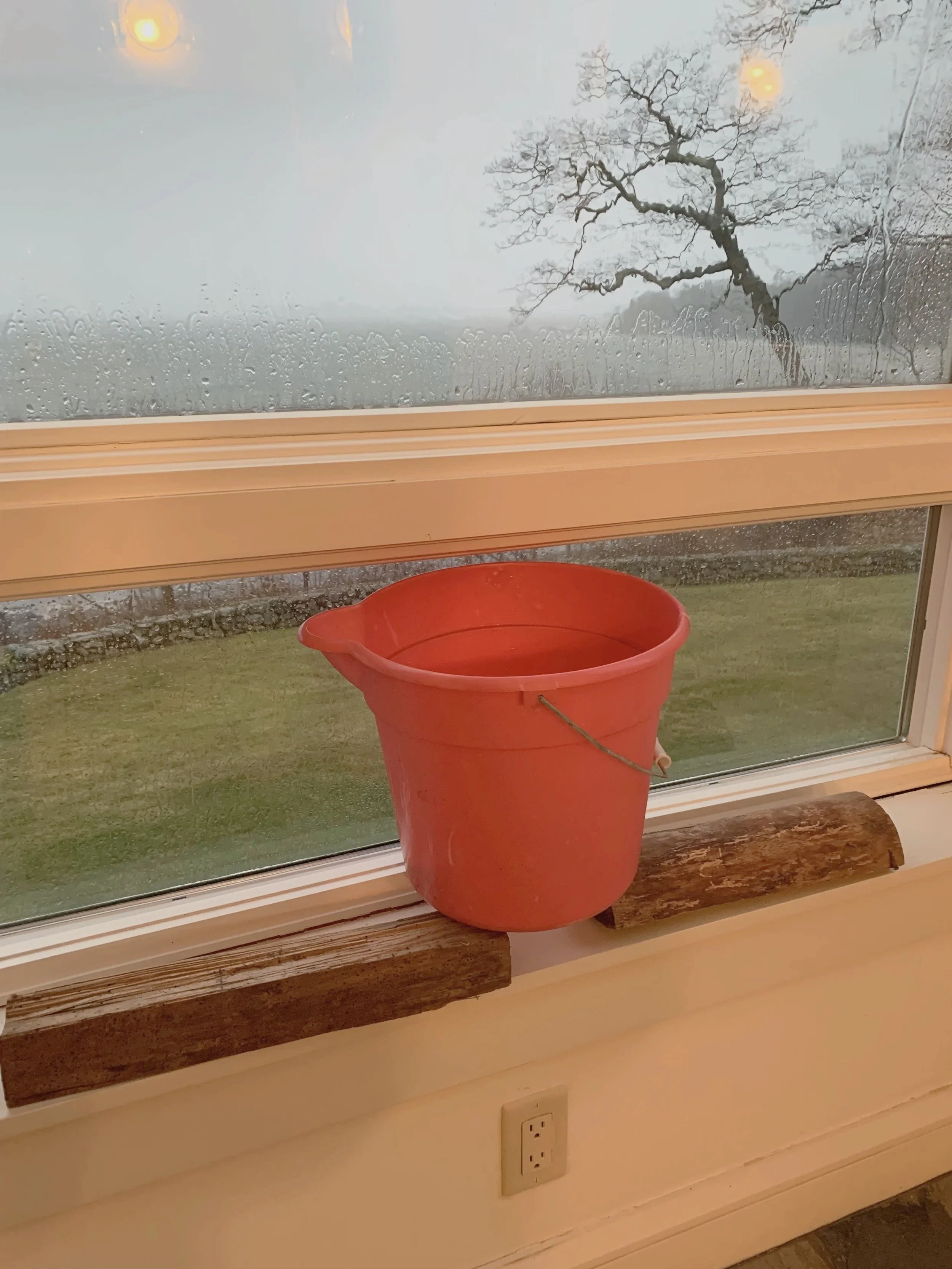A red plastic bucket placed on a wooden window sill inside a house, with two pieces of wood supporting it. Outside the window, there's a rainy landscape with a leafless tree and overcast sky.