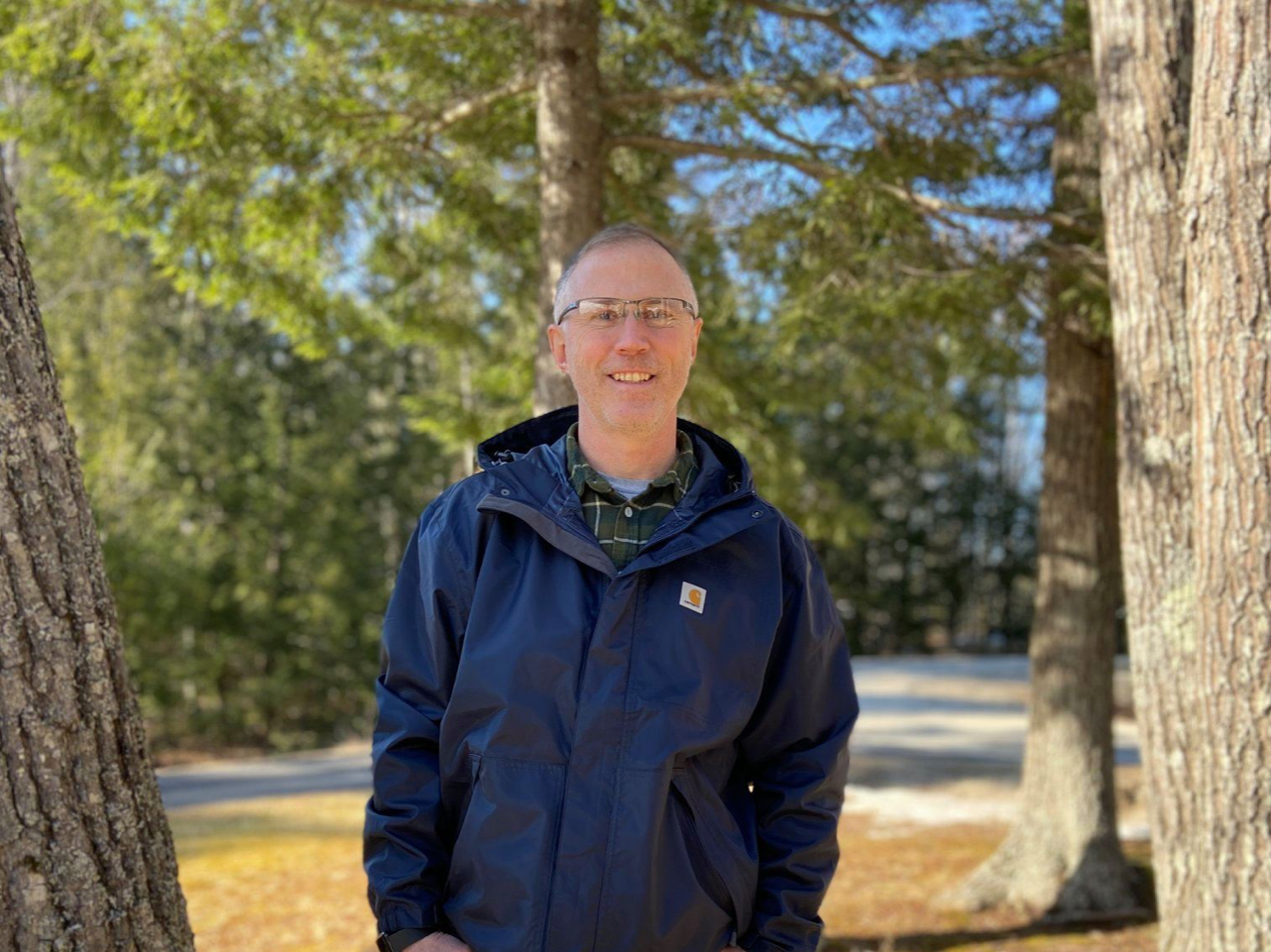 A man smiling outdoors in a green forest with tall trees, wearing glasses, a blue jacket, and a plaid shirt underneath.