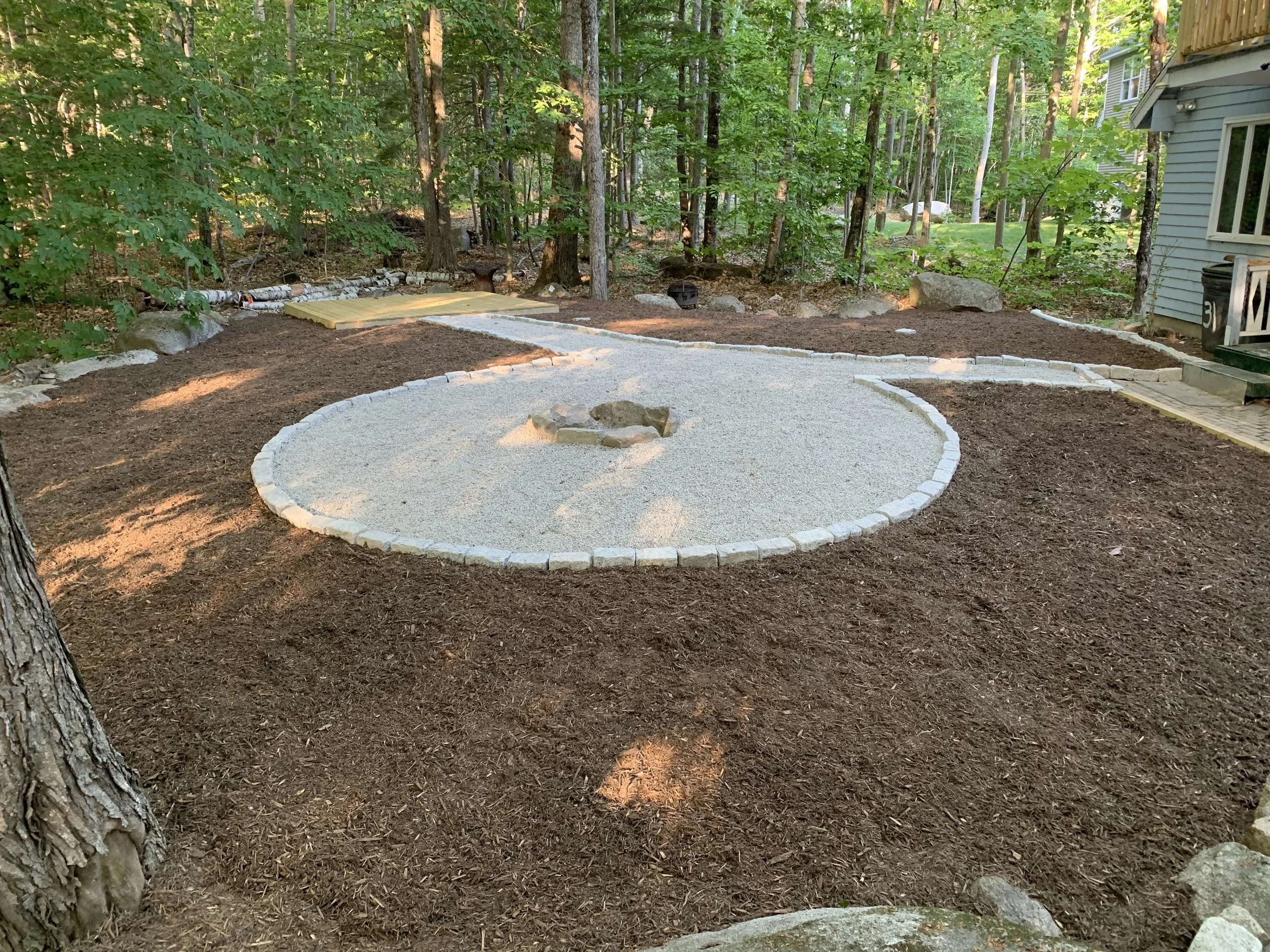 Yard with a circular gravel fire pit surrounded by wood border, mulch, and stepping stones, with trees and a house in the background.