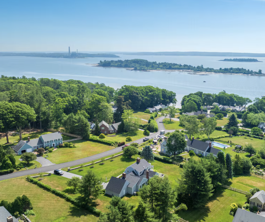 Aerial view of a suburban neighborhood overlooking the ocean, with houses, trees, and a winding road.