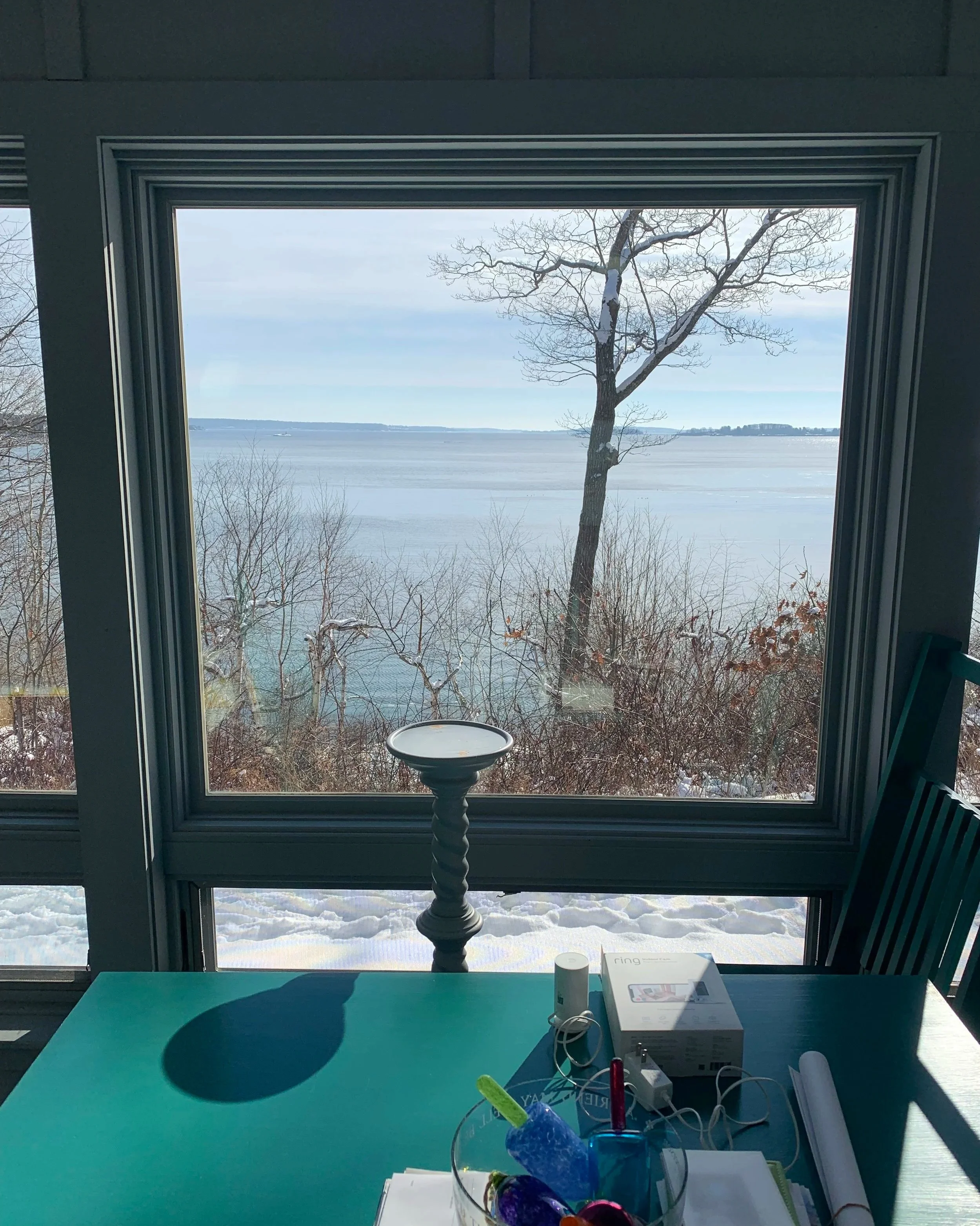 View of a Maine ocean scene through large window from inside a room, with snow on the ground and a leafless tree near the window.