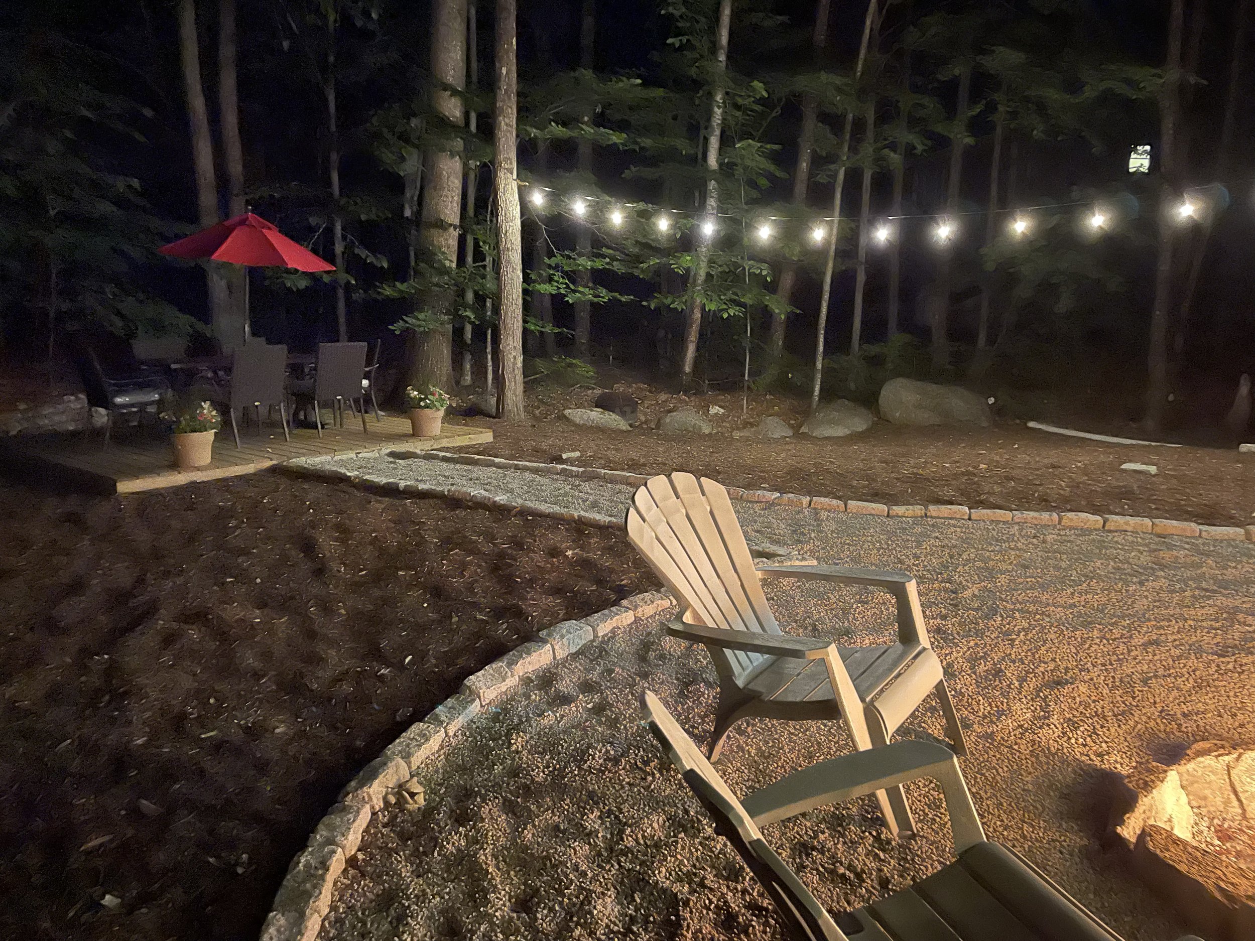 Nighttime outdoor backyard scene with wooden Adirondack chairs, a fire pit, string lights, patio furniture with a red umbrella, and potted plants surrounded by trees and rocks.