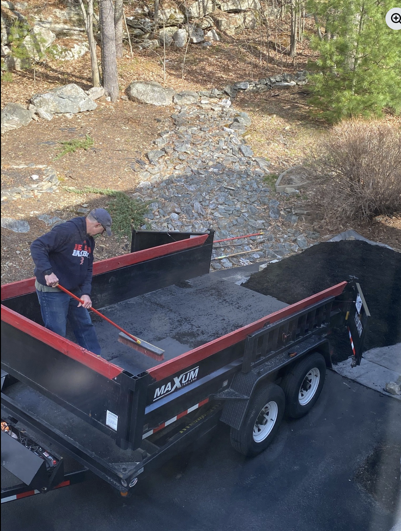 A man pushing mulch off of a black trailer with red edges, parked on a driveway, nearby rocky terrain and trees.