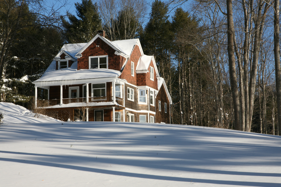 A large, multi-story house with a steep gable roof and a wrap-around porch, covered in snow, surrounded by tall trees on a clear, sunny winter day.