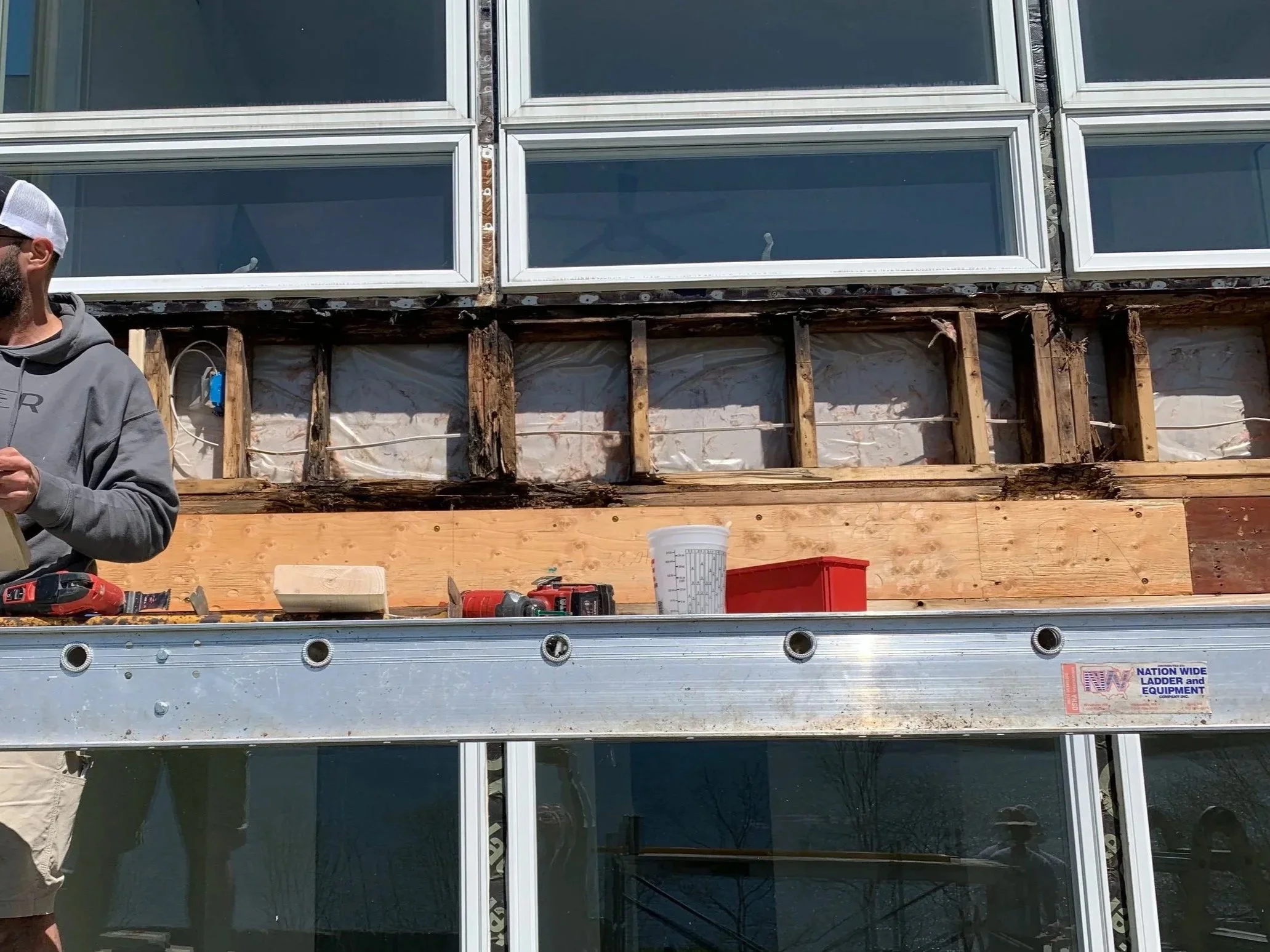 Contractor standing on a scaffold working on building renovation of large glass windows with visible wooden framing and structural supports.