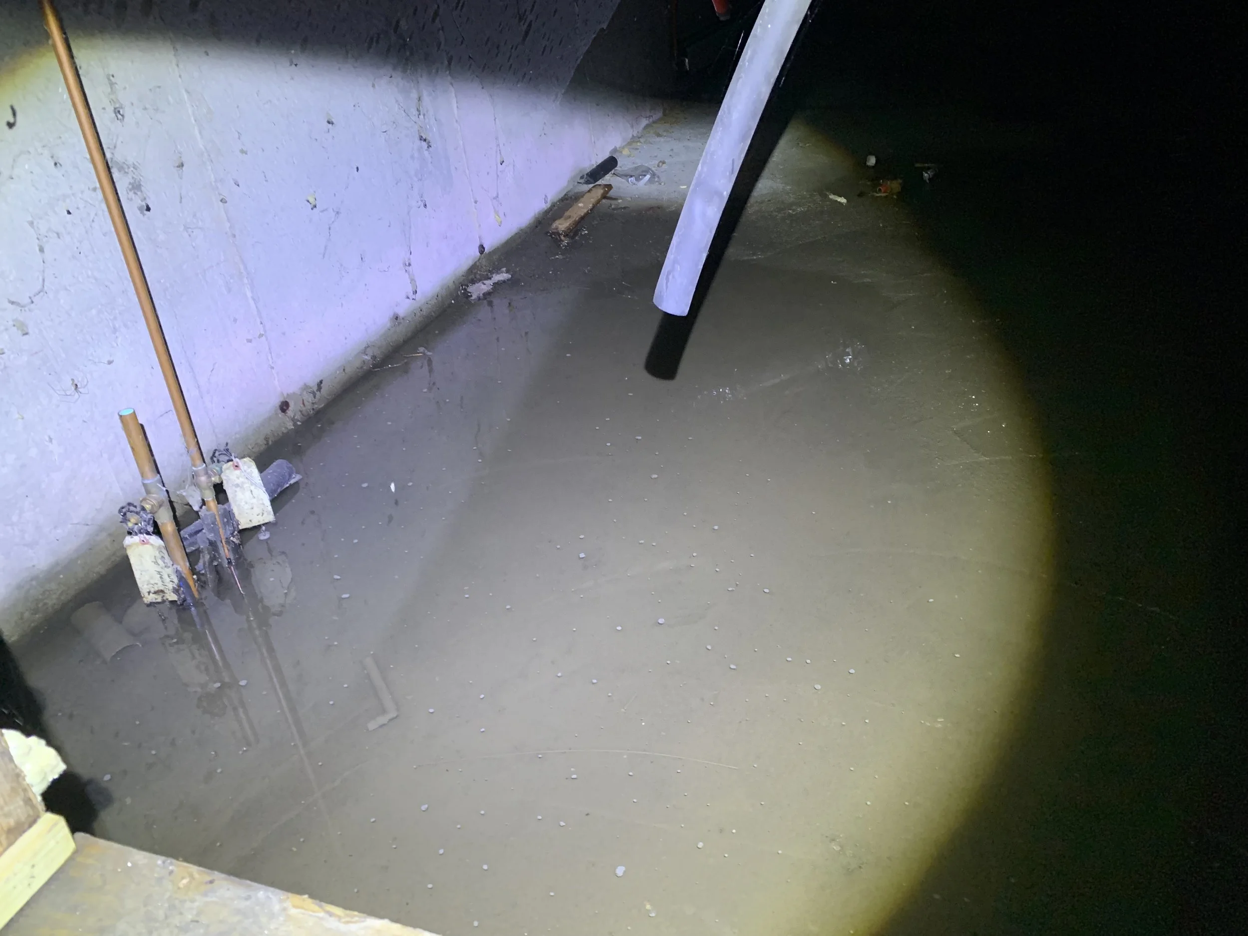 A basement with a concrete floor, two copper pipes with valves, and a recess in the wall as well as a flood of water on the ground.